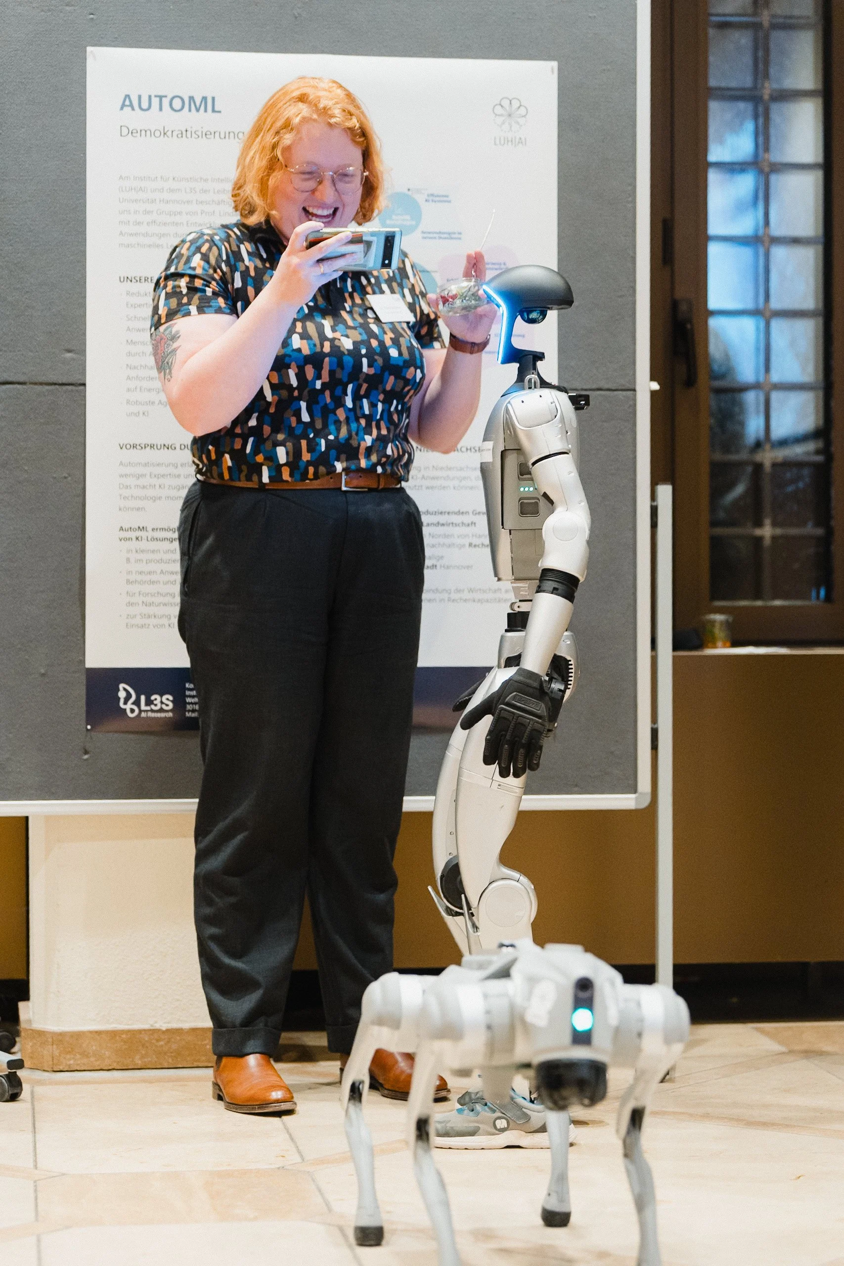 Eine lachende Frau mit roten Haaren, Brille und Tattoo auf dem Arm, steht vor einem Rollstuhlroboter und fotografiert ihn mit ihrem Smartphone. Im Hintergrund ein Poster an einer Wand.