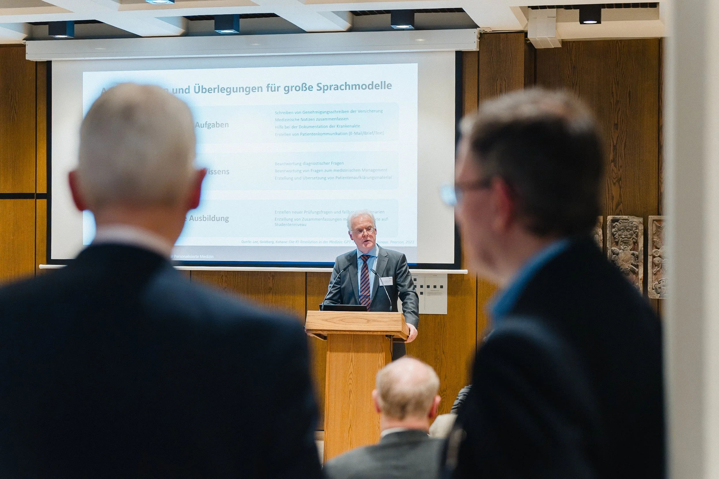 Ein Mann in einem Anzug hält eine Präsentation vor einem Publikum in einem Konferenzraum. Auf der Leinwand sind Textinhalte zu sehen.