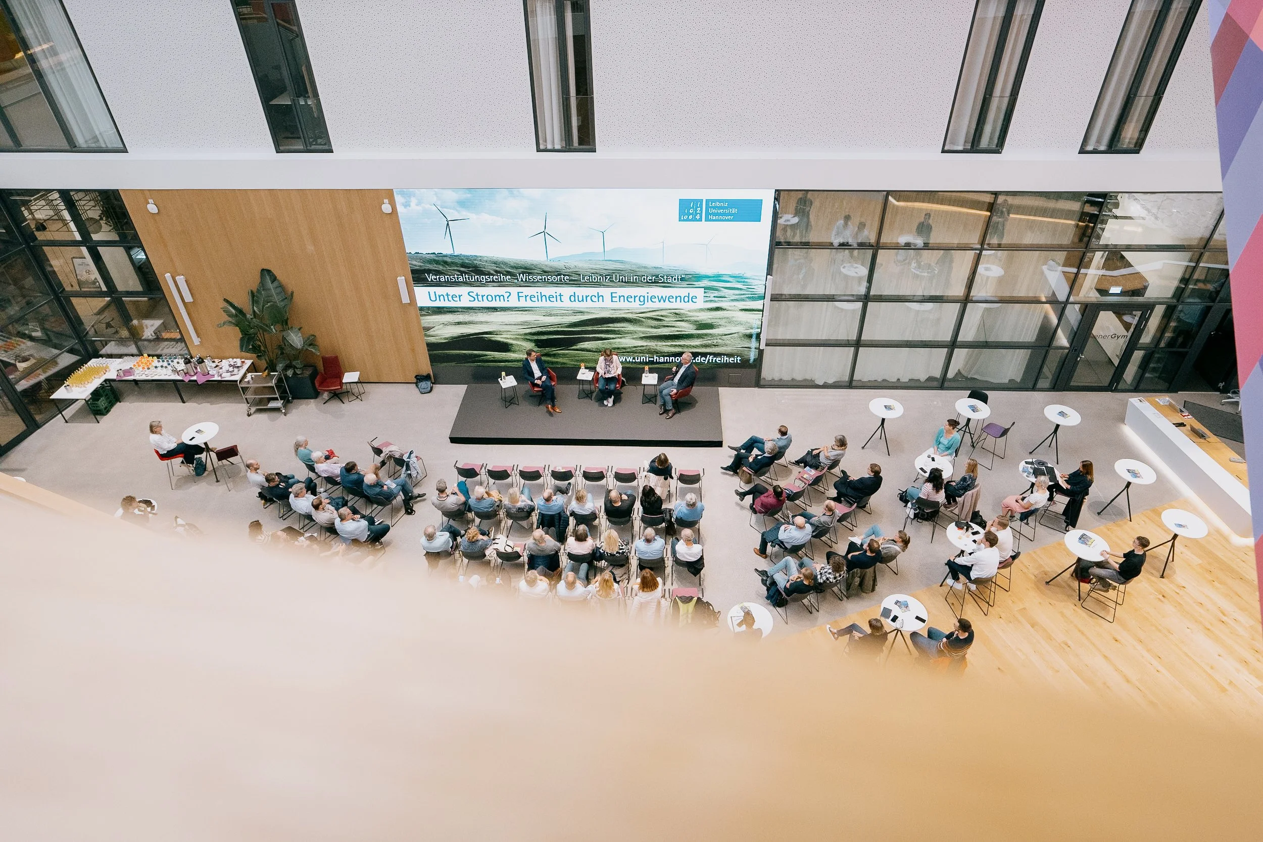 Innenraum eines Konferenzsaals mit einem Publikum, das einem Panel mit drei Sprechern auf der Bühne zuhört. Im Hintergrund eine große digitale Anzeigetafel mit Text und Windenergie-Bild. Rundtische mit Tassen und Papieren sind im Raum verteilt.