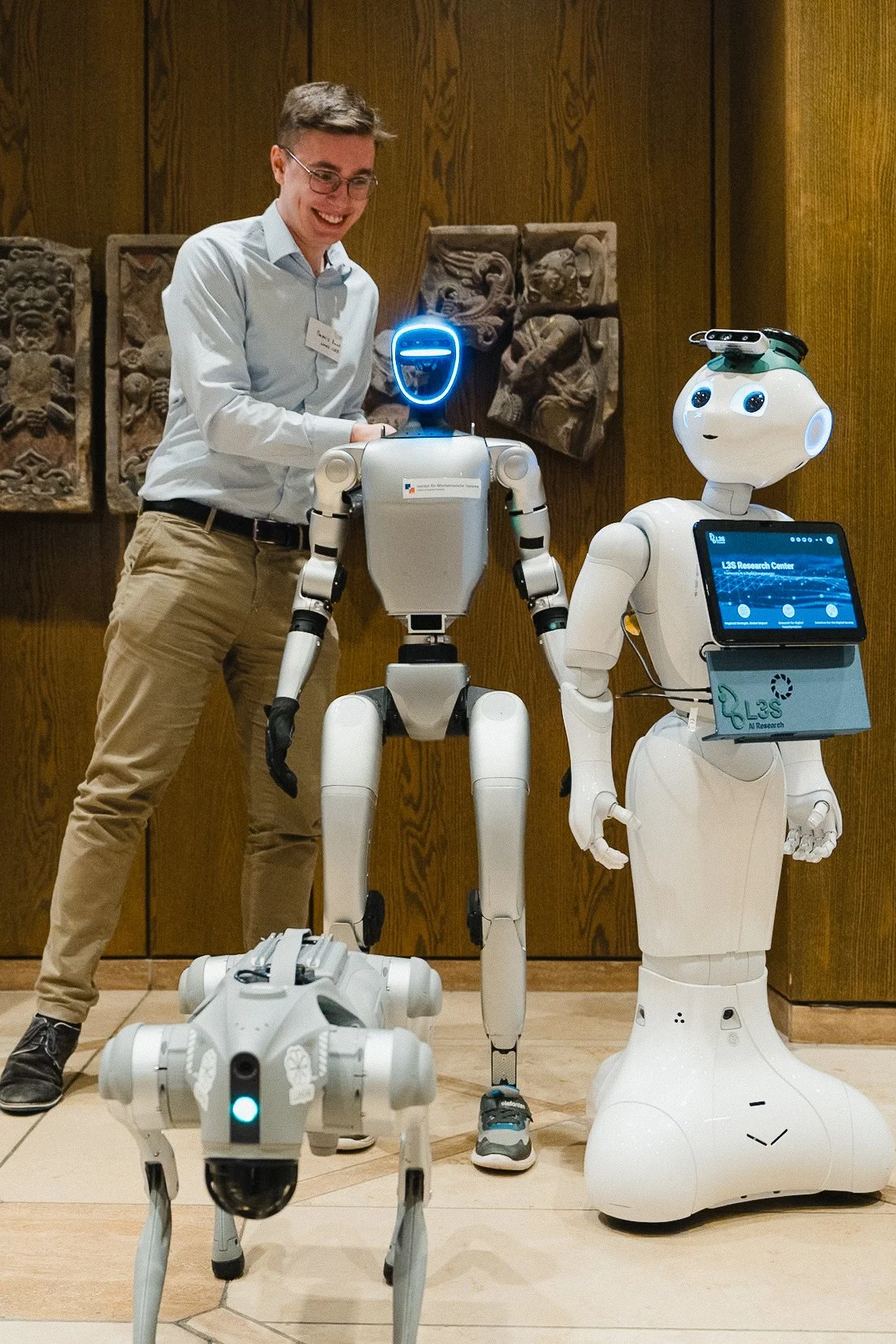 Ein Mann im Hemd und Jeans neben drei Robotern in einem Raum mit Holzwänden und Wanddekorationen.