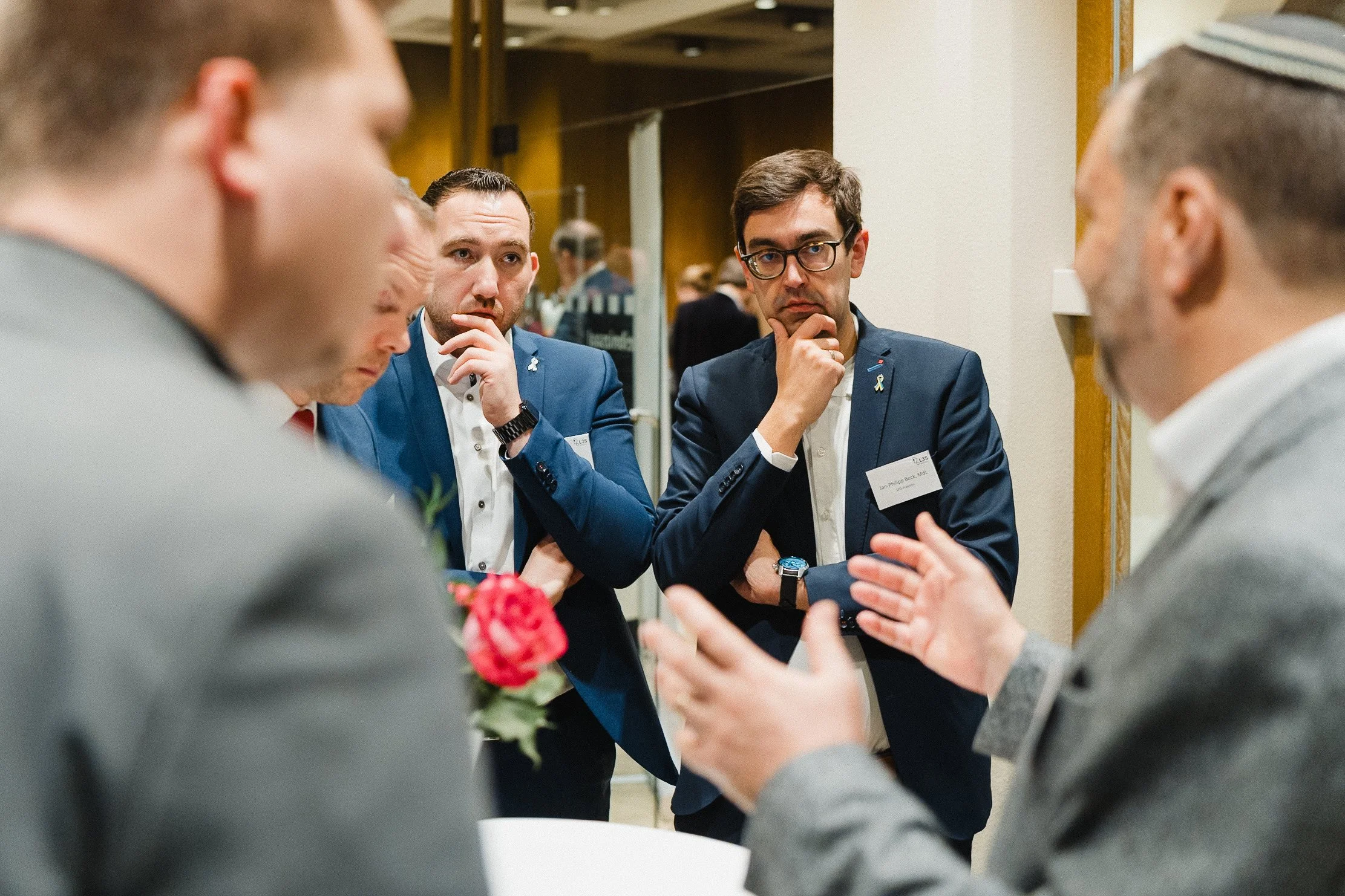 Vier Männer in Anzügen führen eine Diskussion bei einer Veranstaltung, ein Mann auf der rechten Seite erklärt etwas, während die anderen Männer aufmerksam zuhören.