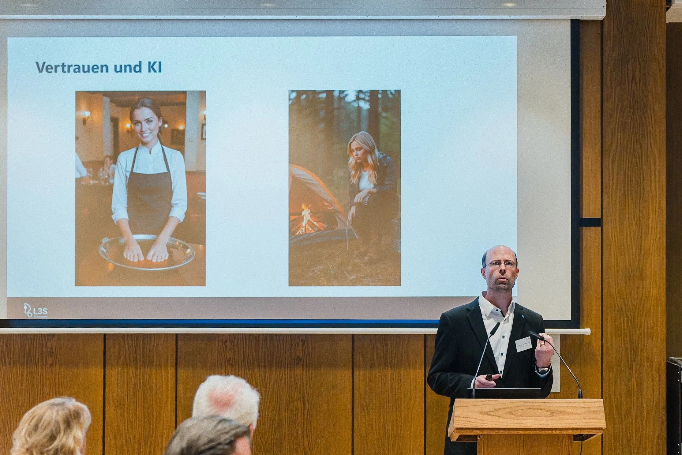 Ein Mann präsentiert eine PowerPoint-Folie mit dem Titel 'Vertrauen und KI'. Auf der Folie sind zwei Fotos: links eine Frau in einem Restaurant, rechts eine Frau beim Zelten im Wald.