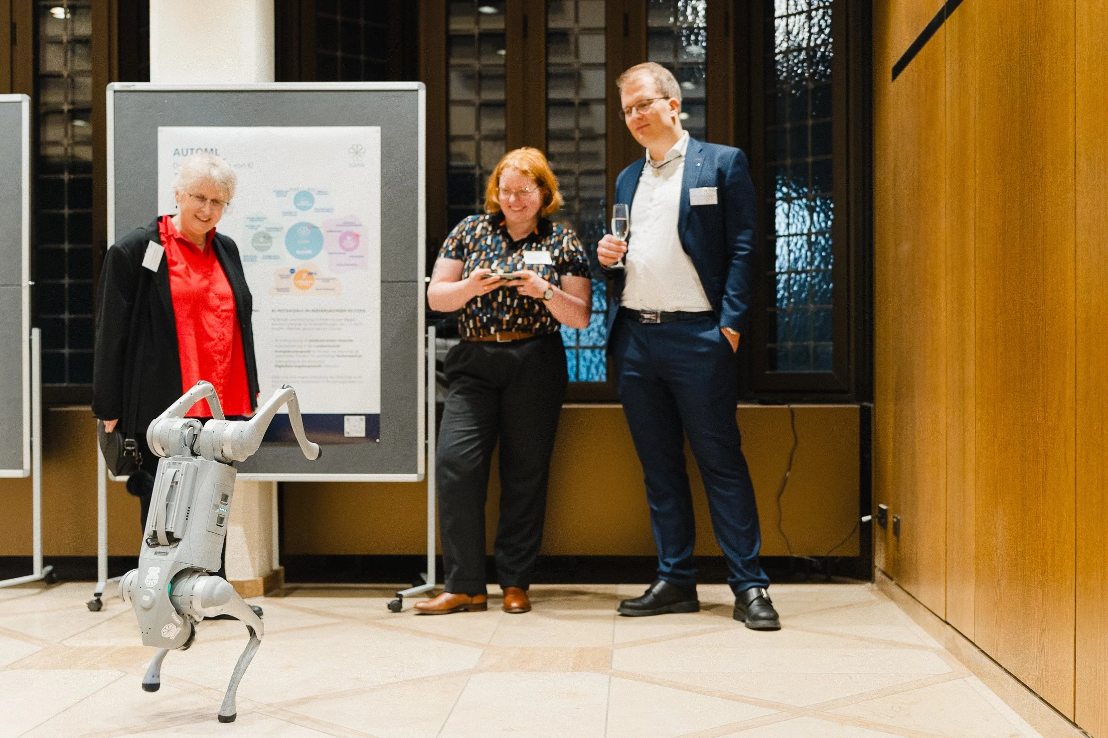 Drei Menschen stehen in einem Raum mit Holzwänden und großen Fenstern, eine Frau, ein Mann und eine ältere Frau. Der Mann hält ein Glas Sekt, die Frauen schauen auf ein Smartphone. Vor ihnen steht ein humanoider Roboter auf drei Beinen.