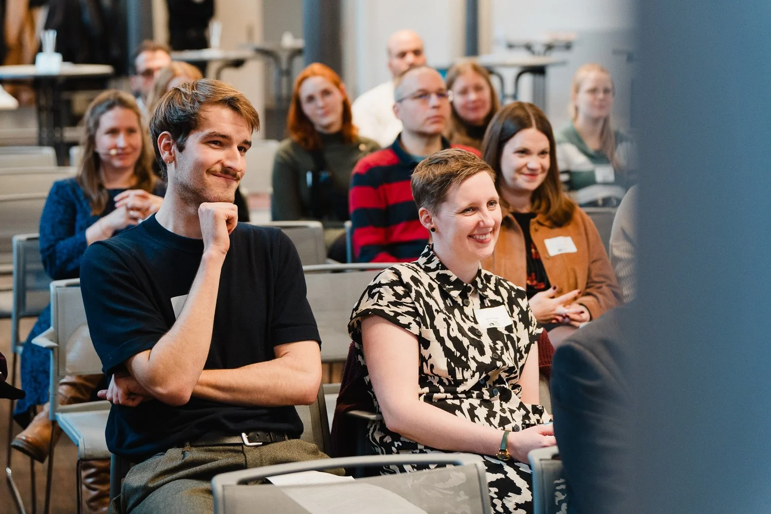 Menschen sitzen in einem Saal bei einer Konferenz, einige lachen, andere schauen interessiert.