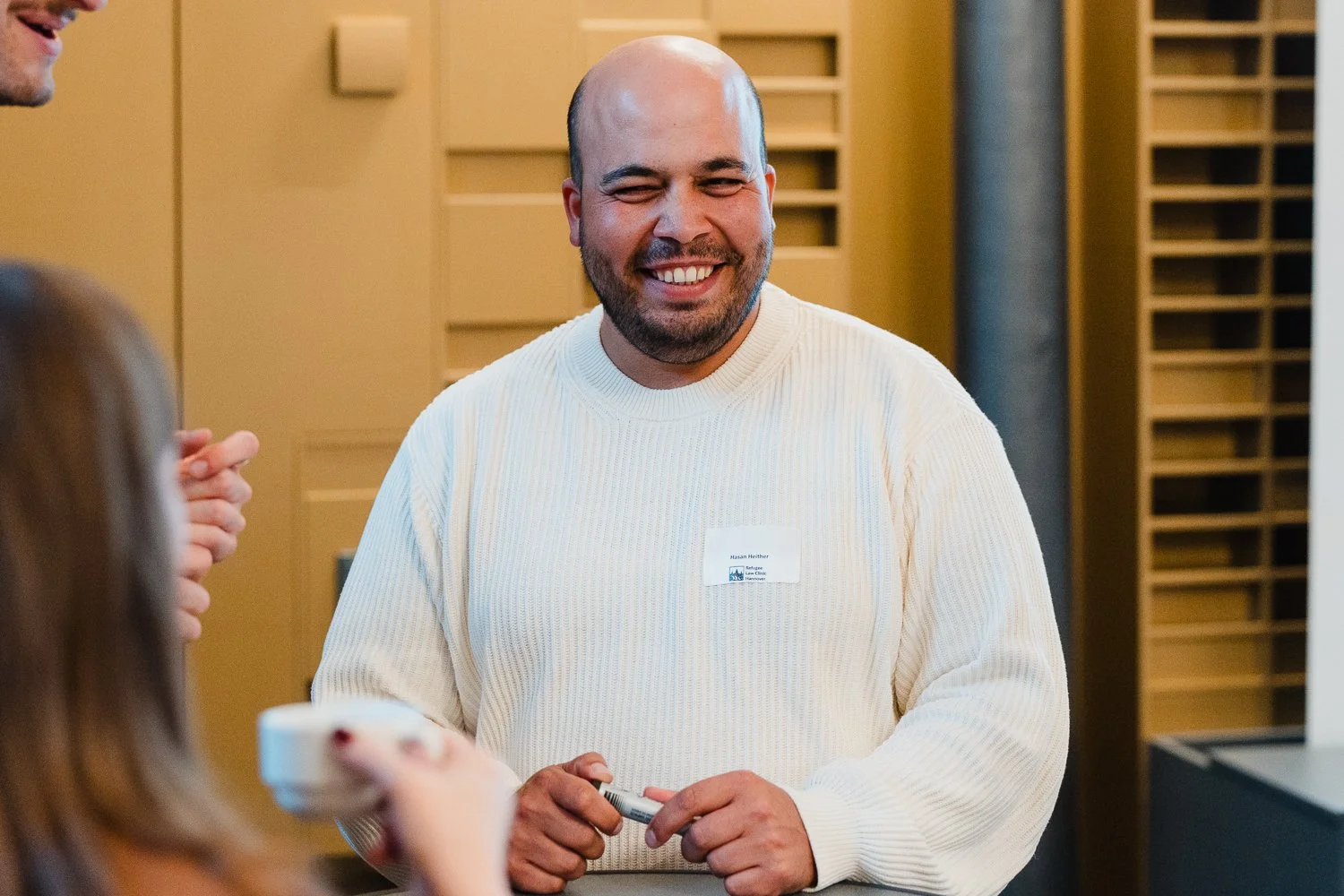 Ein lachender Mann mit Glatze in einem weißen Pullover, der an einem Gespräch teilnimmt, während andere Personen im Vordergrund sind.