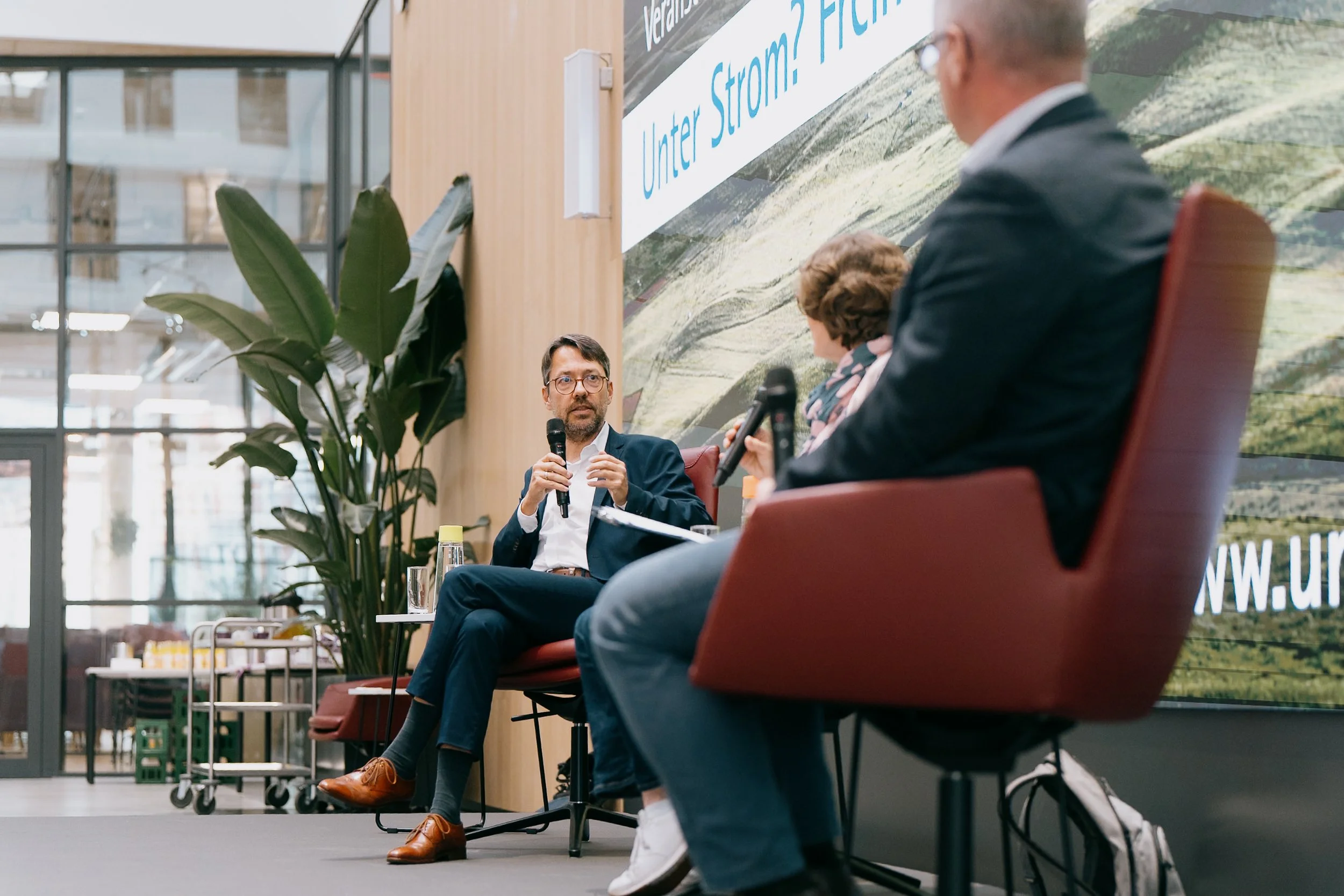 Drei Menschen in einer Diskussionsrunde, die in einem modernen Raum sitzen. Der Mann in der Mitte hält ein Mikrofon und spricht, der andere Mann und die Frau hören zu. Im Hintergrund ist eine große Wand mit einem Text und Fenster mit Blick nach drauß