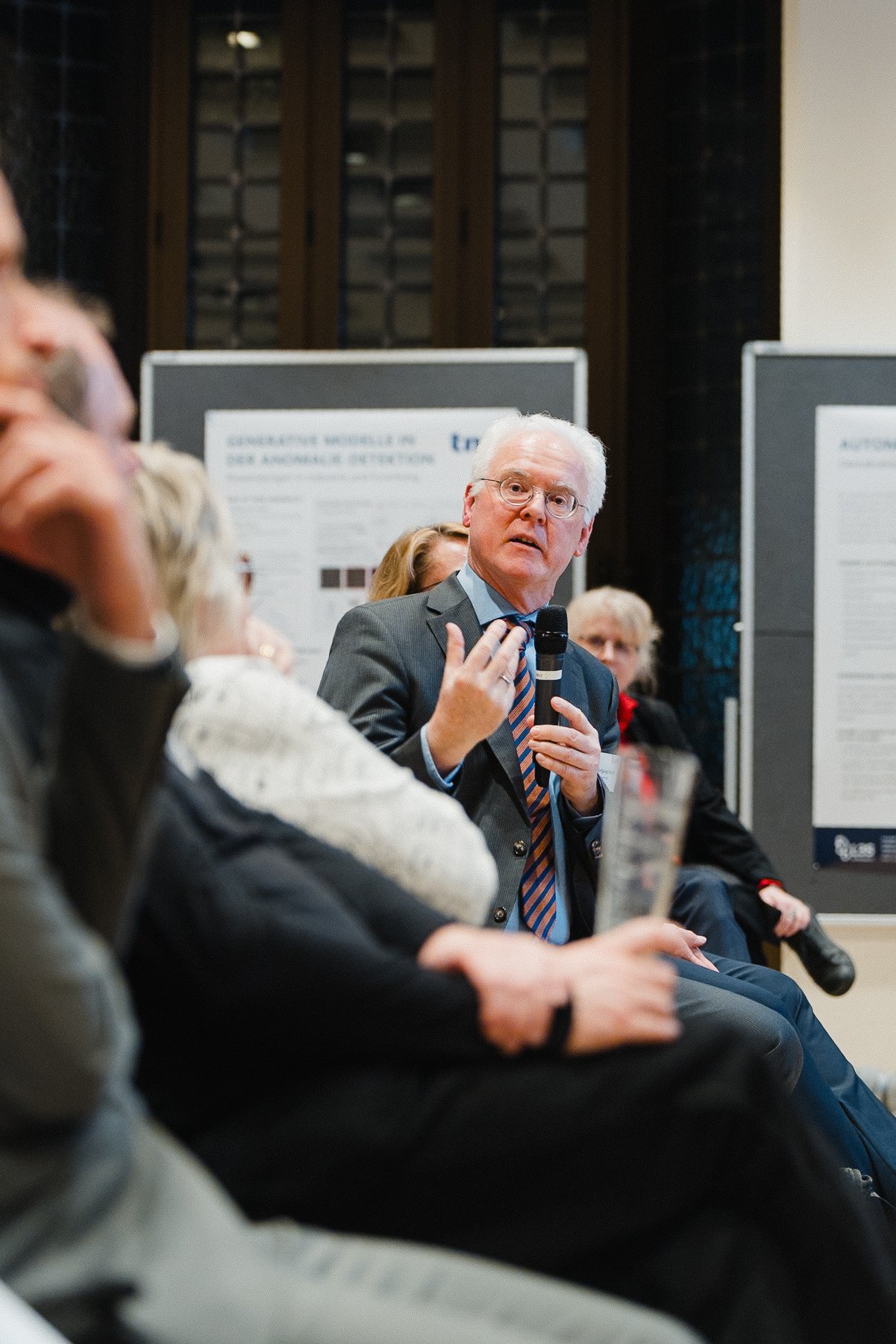 Ein älterer Mann in Anzug und Krawatte hält ein Mikrofon in der Hand und spricht auf einer Konferenz, während die Zuhörer aufmerksam zuhören.