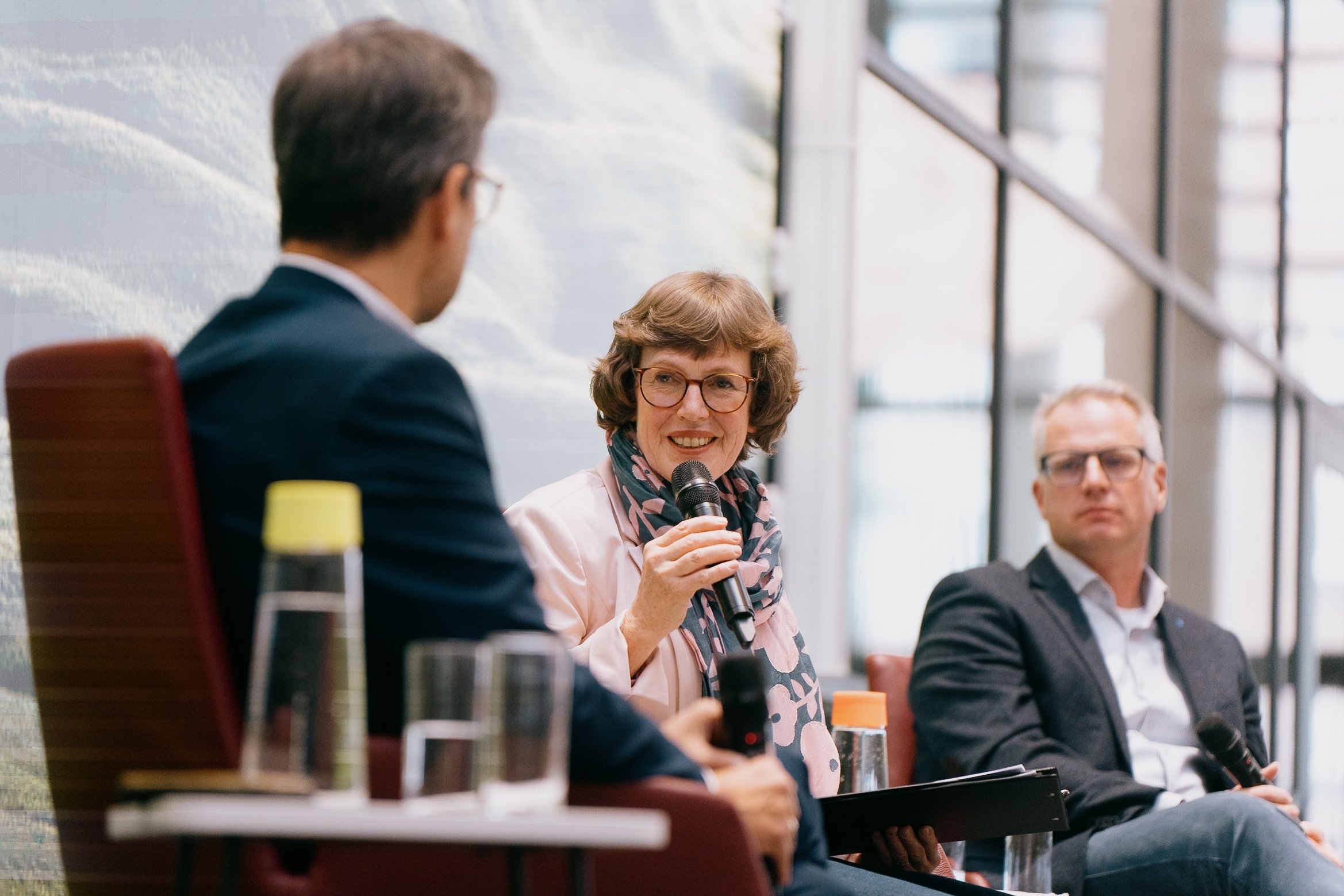 Drei Menschen bei einer Podiumsdiskussion in einem modernen Raum mit großen Fenstern. Die Frau in der Mitte spricht mit einem Mikrofon, während die beiden Männer zuhören.
