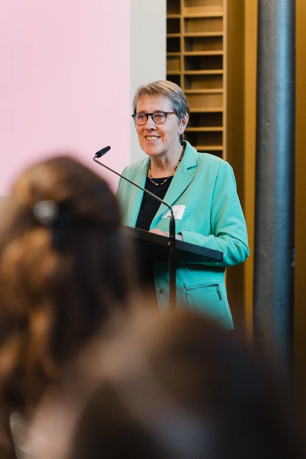 Eine Frau steht an einem Rednerpult und spricht zu einem Publikum, hinter ihr ist eine rosa Wand und ein Bücherregal mit Fächern zu sehen.