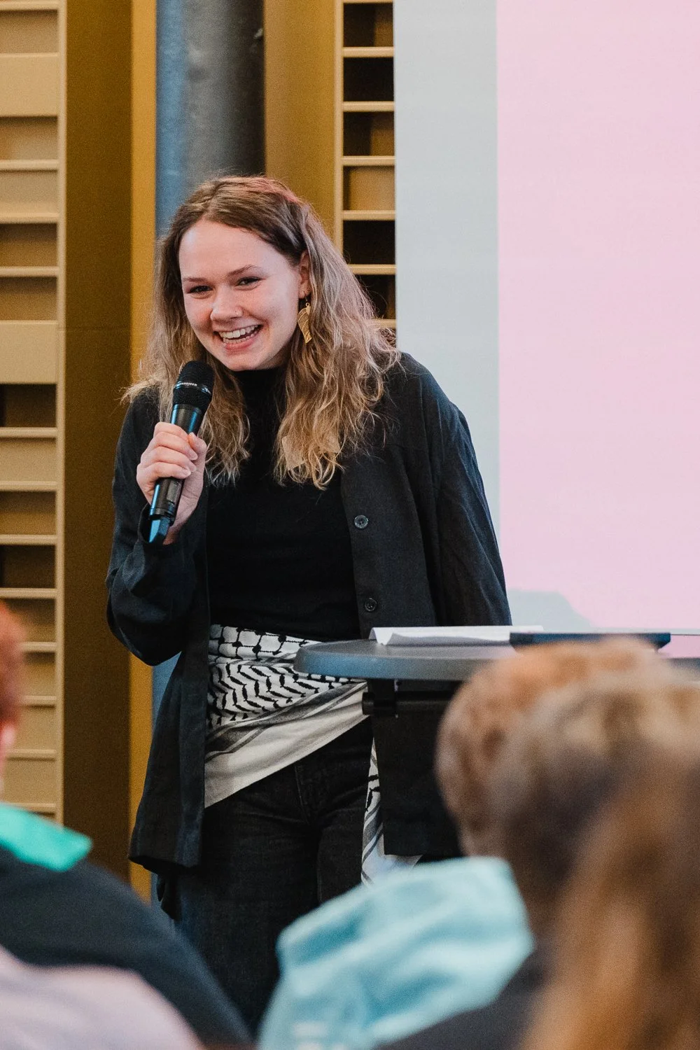 Junge Frau mit Mikrofon, spricht vor Publikum in einem modernen Raum.