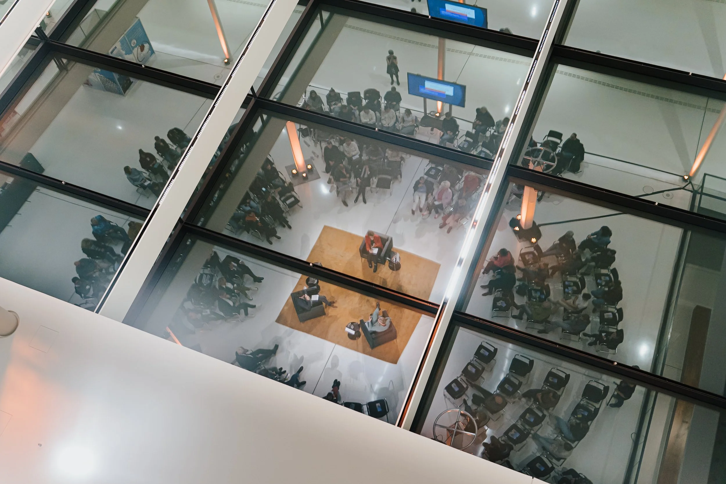 Blick von oben auf die mehrere Etagen eines modernen Gebäudes mit mehreren Warteschlangen, Wartebereichen, Sitzgruppen, einem Service-Schalter, Wartemarken und Menschen, die auf den Aufzug warten.