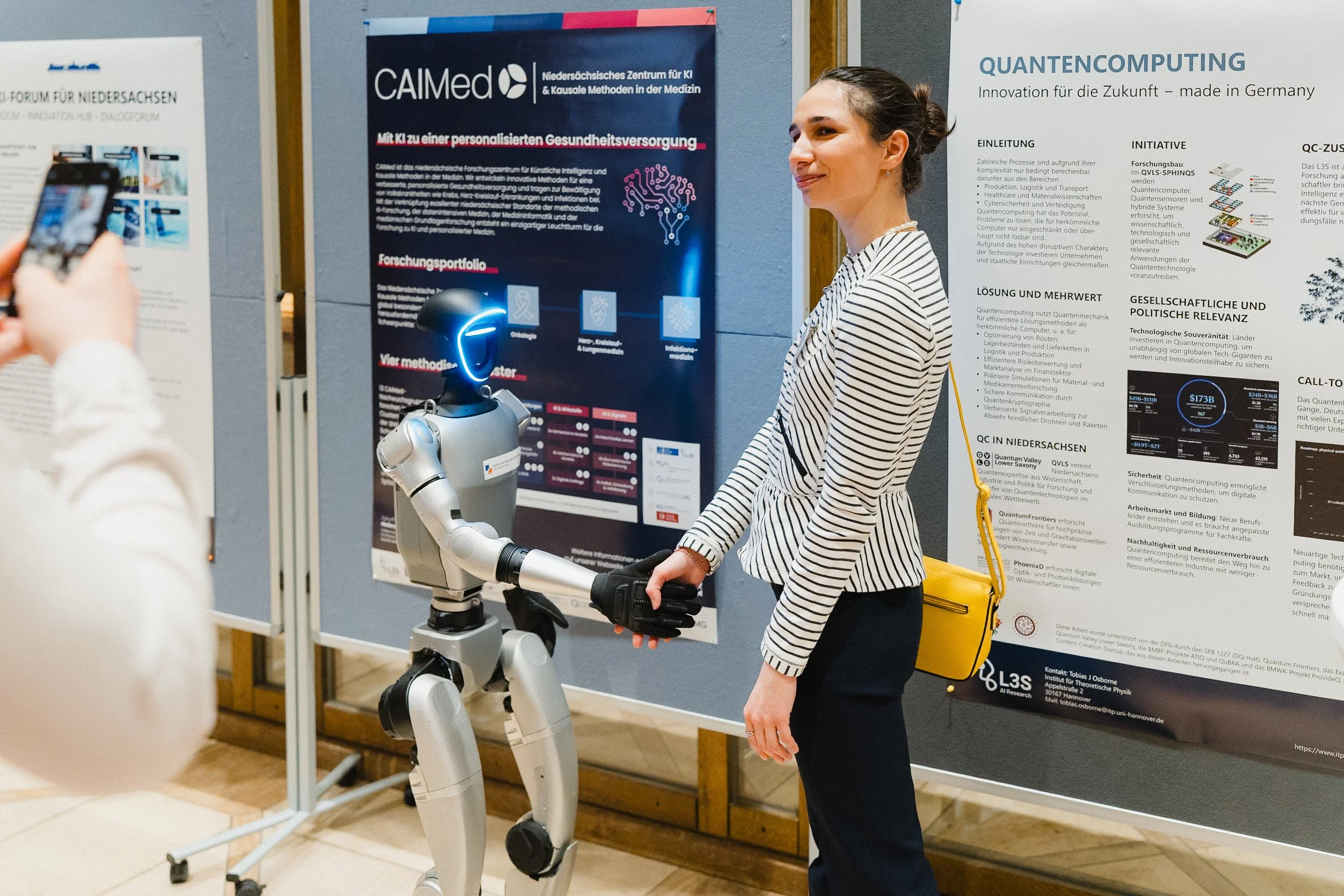 Eine Frau schüttelt die Hand eines Roboters bei einer Konferenz oder Ausstellung, im Hintergrund Poster über Quantencomputing und KI in der Medizin.