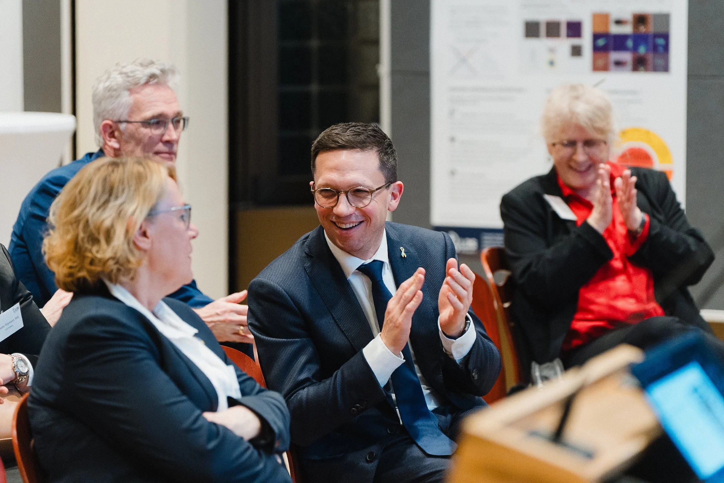 Menschen bei einer Veranstaltung, lachen und klatschen, im Hintergrund Poster mit Grafiken.
