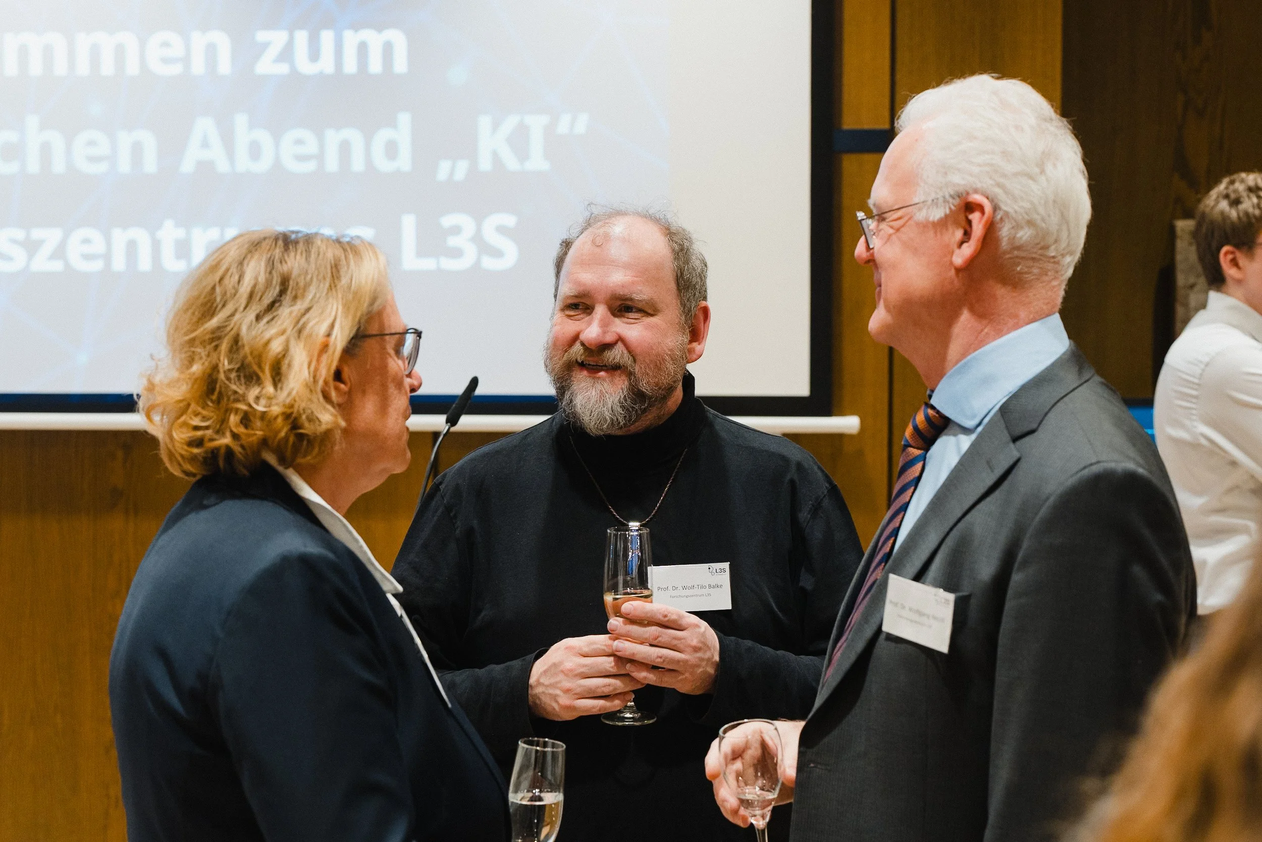 Drei Menschen im Gespräch bei einer Abendveranstaltung, alle halten Gläser mit Getränken, im Hintergrund ein Präsentation mit deutschem Text auf einem Bildschirm.