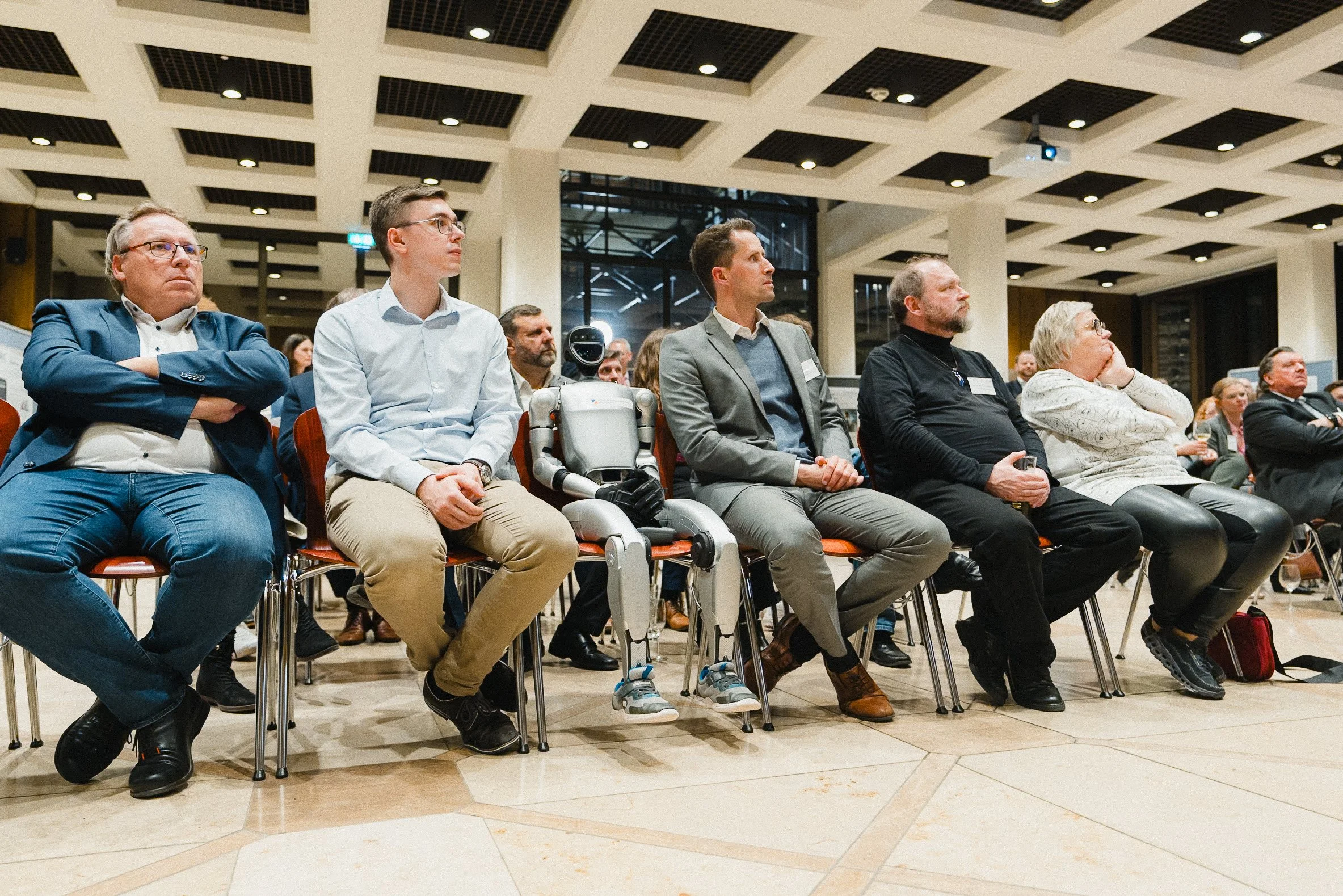 Menschen sitzen in einem Konzertsaal während einer Konferenz, darunter ein humanoider Roboter.
