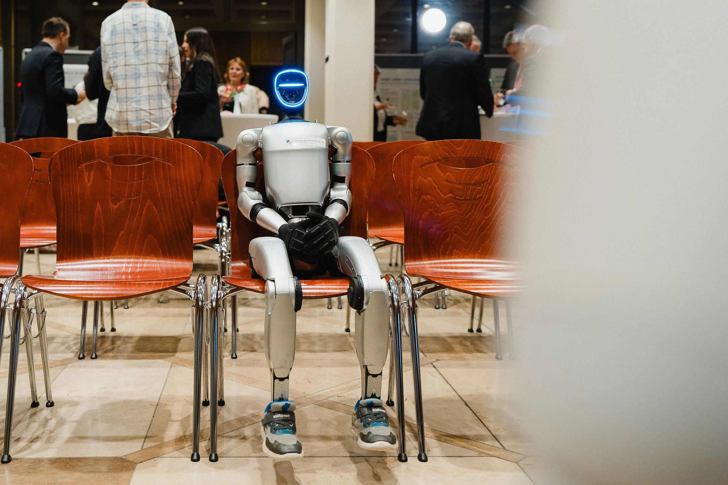 Ein humanoider Roboter sitzt auf einem Stuhl in einem Wartebereich in einem Gebäude, im Hintergrund sind mehrere Menschen, die sich unterhalten und warten.