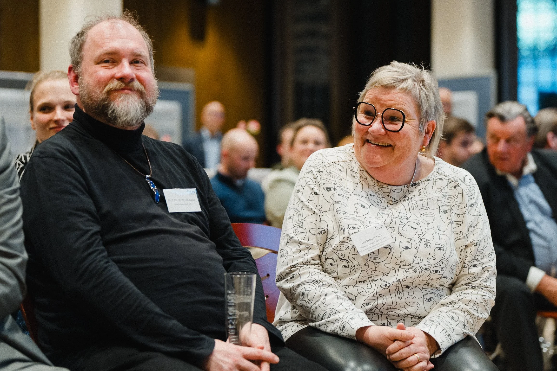 Menschen sitzen in einem Raum, einer hält ein Glas. Im Hintergrund weitere Personen, einige schauen nach vorne, andere reden. Die Stimmung ist freundschaftlich.