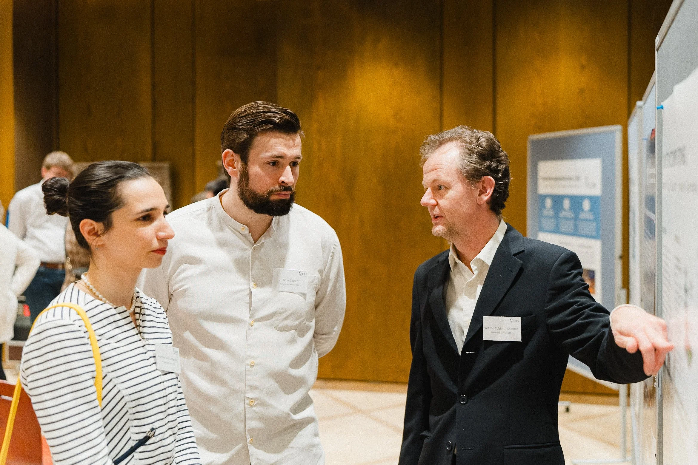 Drei Menschen bei einer wissenschaftlichen Präsentation in einem Raum mit Holzvertäfelung, ein Mann erklärt einen Forschungsstand einem jungen Mann und einer jungen Frau.