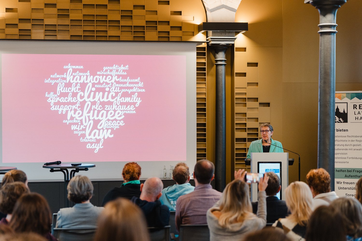Eine Frau spricht an einem Rednerpult vor einer Gruppe von Menschen in einem großen Saal. Auf der großen Leinwand hinter ihr ist eine Wortwolke in Herzform mit verschiedenen deutschen Begriffen zu sehen.