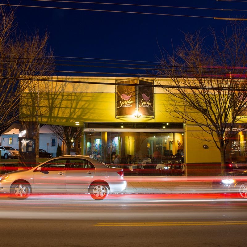 The image shows a yellow building with a restaurant named 'Saico' on the front. The restaurant has large glass windows revealing the interior. There are cars on the street in front, with light streaks indicating long exposure photography, and bare tr
