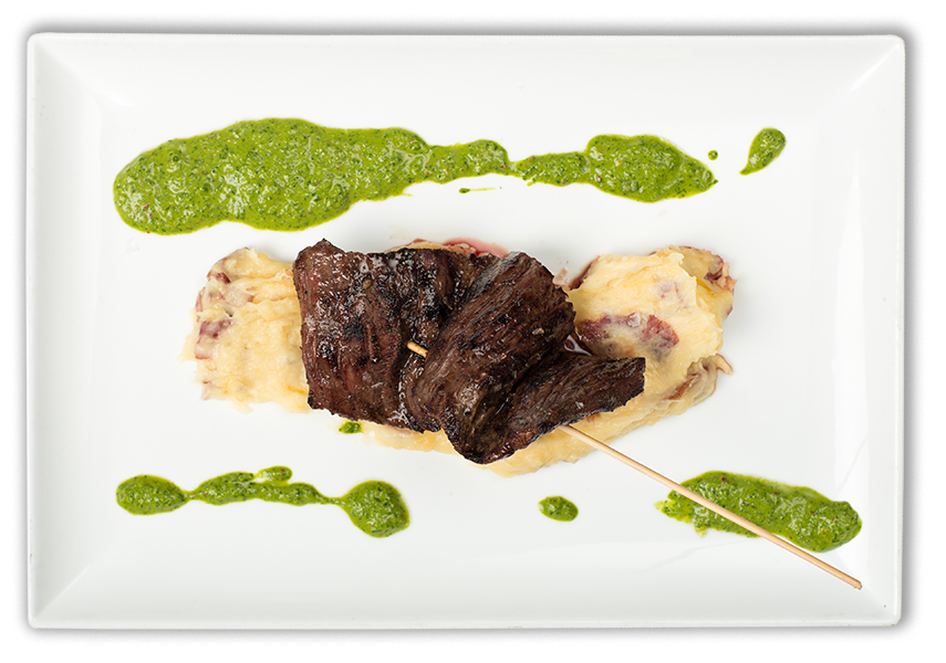 A gourmet dish featuring roasted meat on a bed of mashed potatoes, garnished with green herb sauce, served on a white rectangular plate.