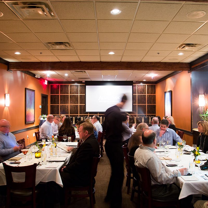 People seated at tables in a restaurant or banquet room, waiting for an event or presentation, with a large screen at the front of the room.