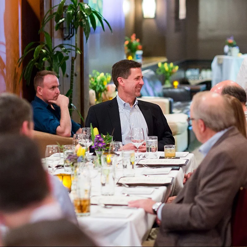 People sitting at a decorated dining table, one man smiling and engaging in conversation, surrounded by glasses and floral centerpieces, in a well-lit restaurant or banquet hall.