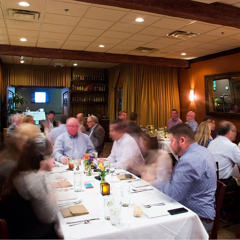 People seated at a long dinner table in a warmly lit restaurant or banquet hall, with some motion blur indicating movement.