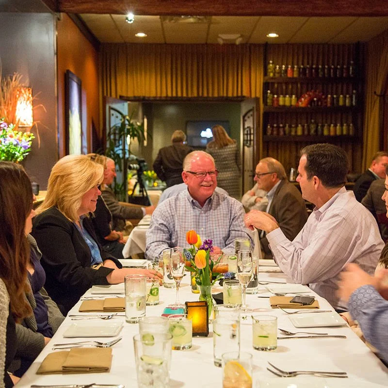 Group of people dining and talking at a long table in a warmly lit restaurant with floral centerpieces and glasses of water and drinks.