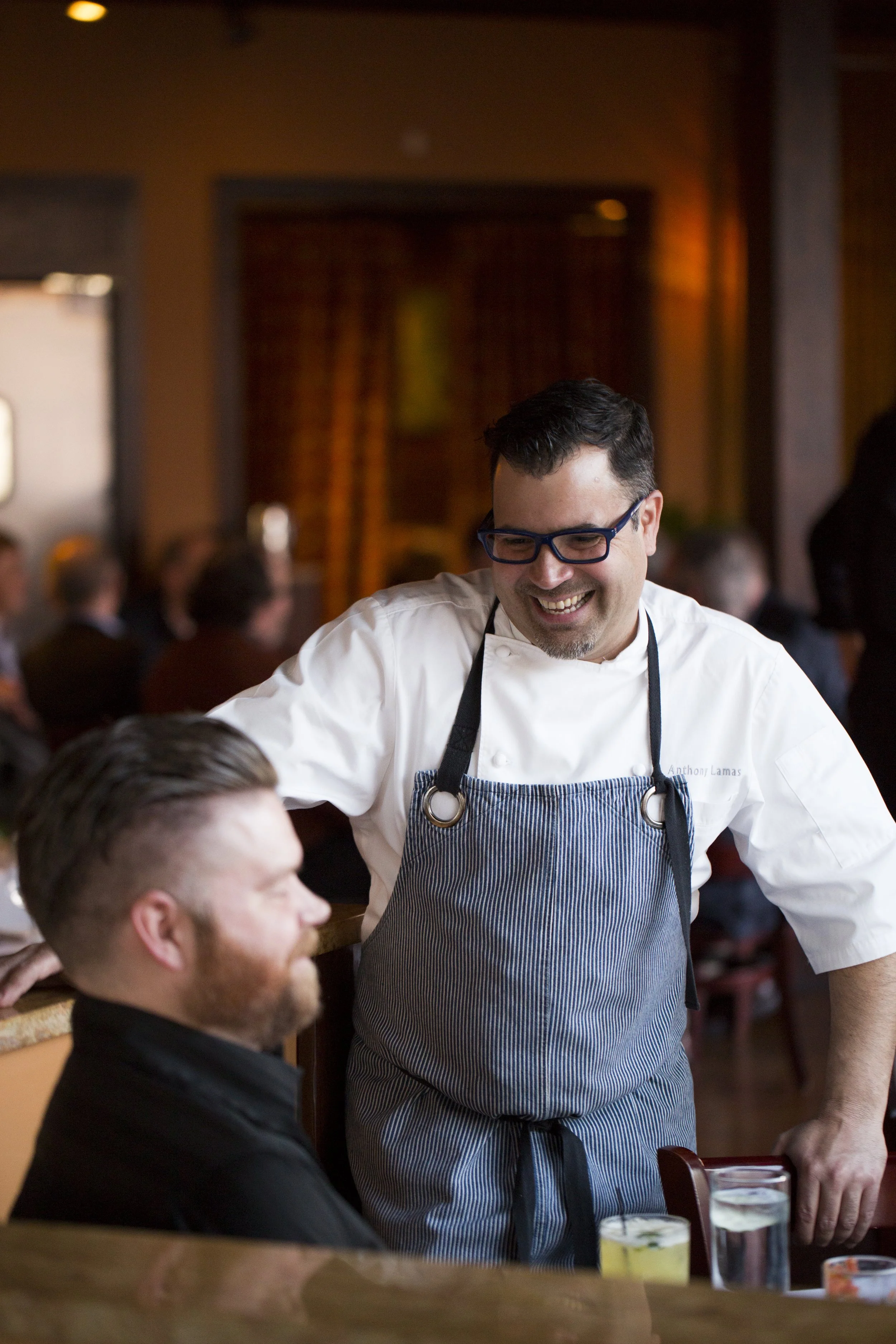 A chef with glasses and a striped apron smiling and talking to a seated customer in a restaurant filled with people in the background.