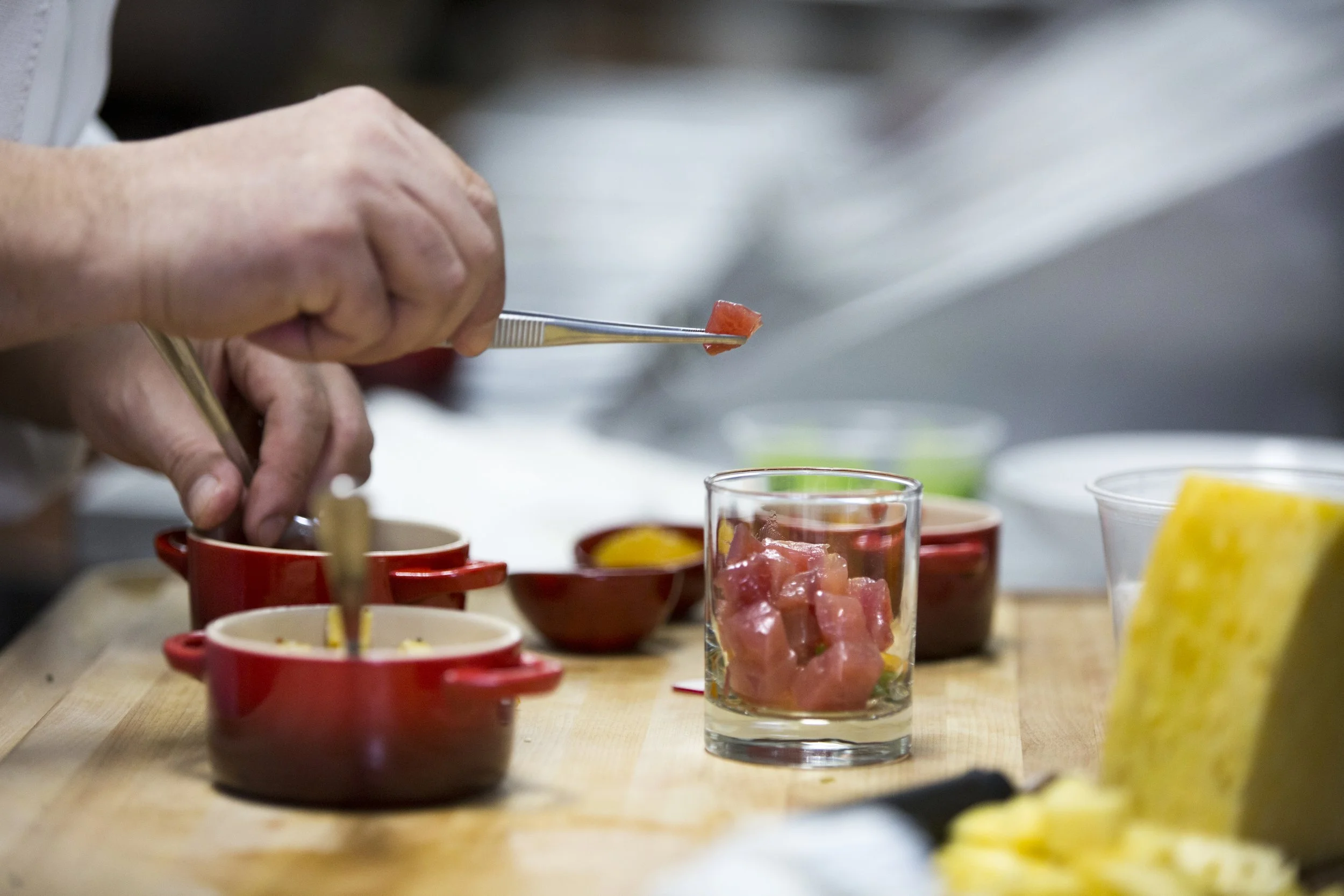 A chef preparing diced tuna for a dish in a professional kitchen, with various bowls and ingredients on a wooden counter.