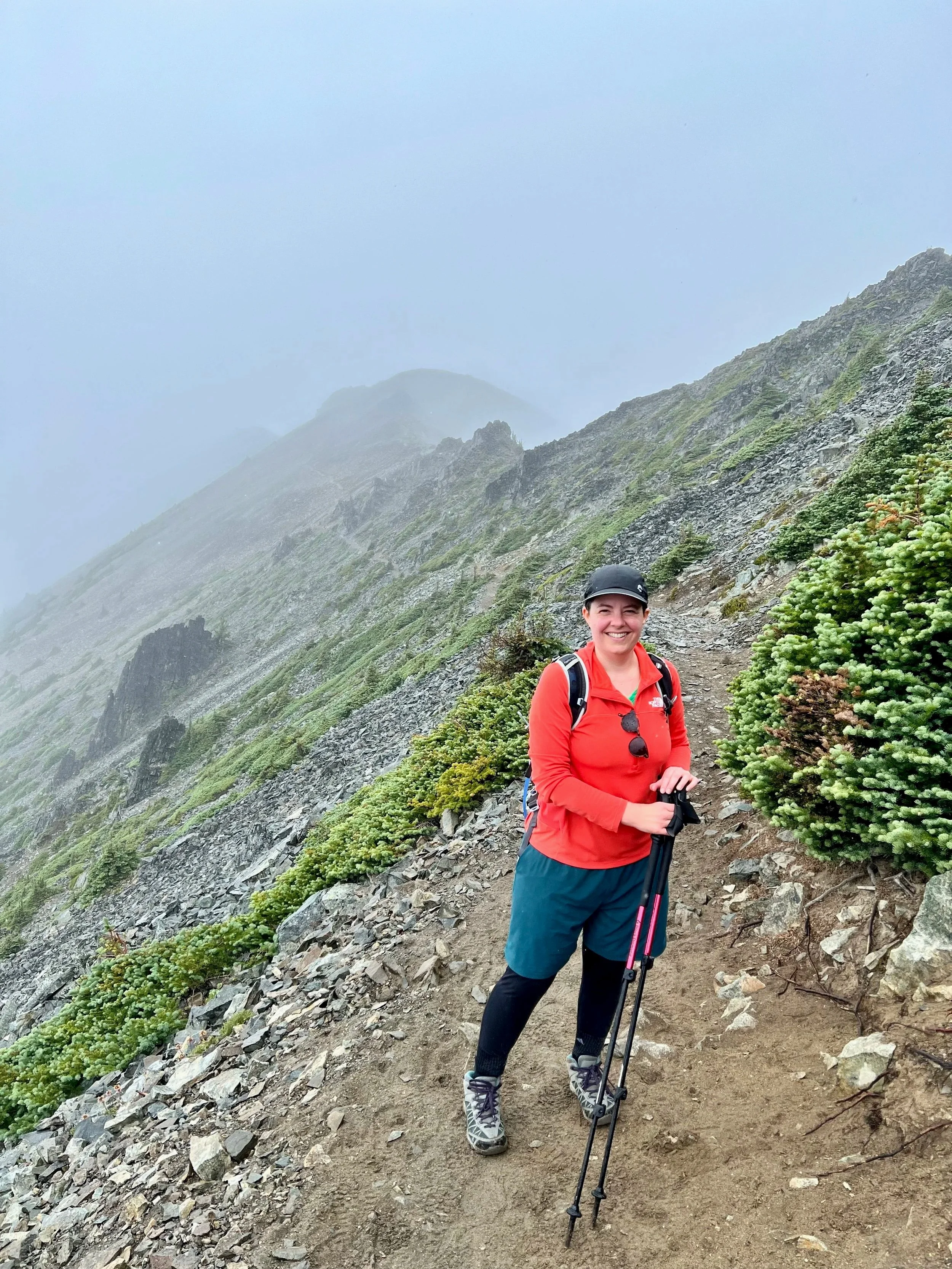A woman hiking on a mountain trail with rocky terrain and green plants, wearing a red jacket, black cap, hiking shoes, and holding trekking poles, smiling with a misty mountain landscape in the background.