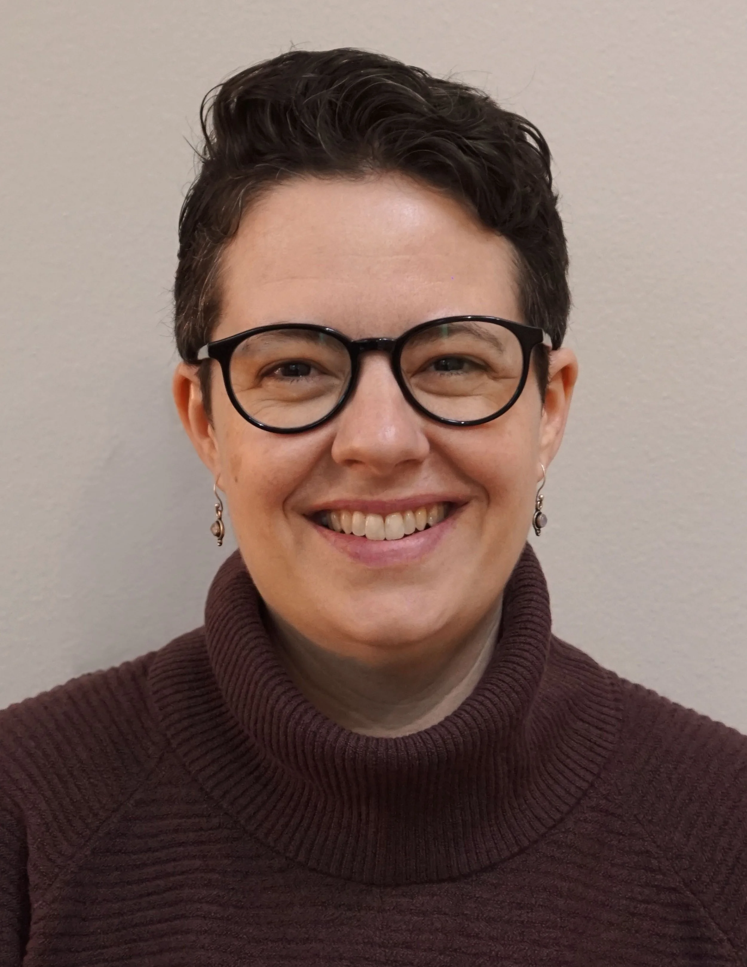 Close-up of a person smiling, wearing glasses and a burgundy sweater, against a plain light background.
