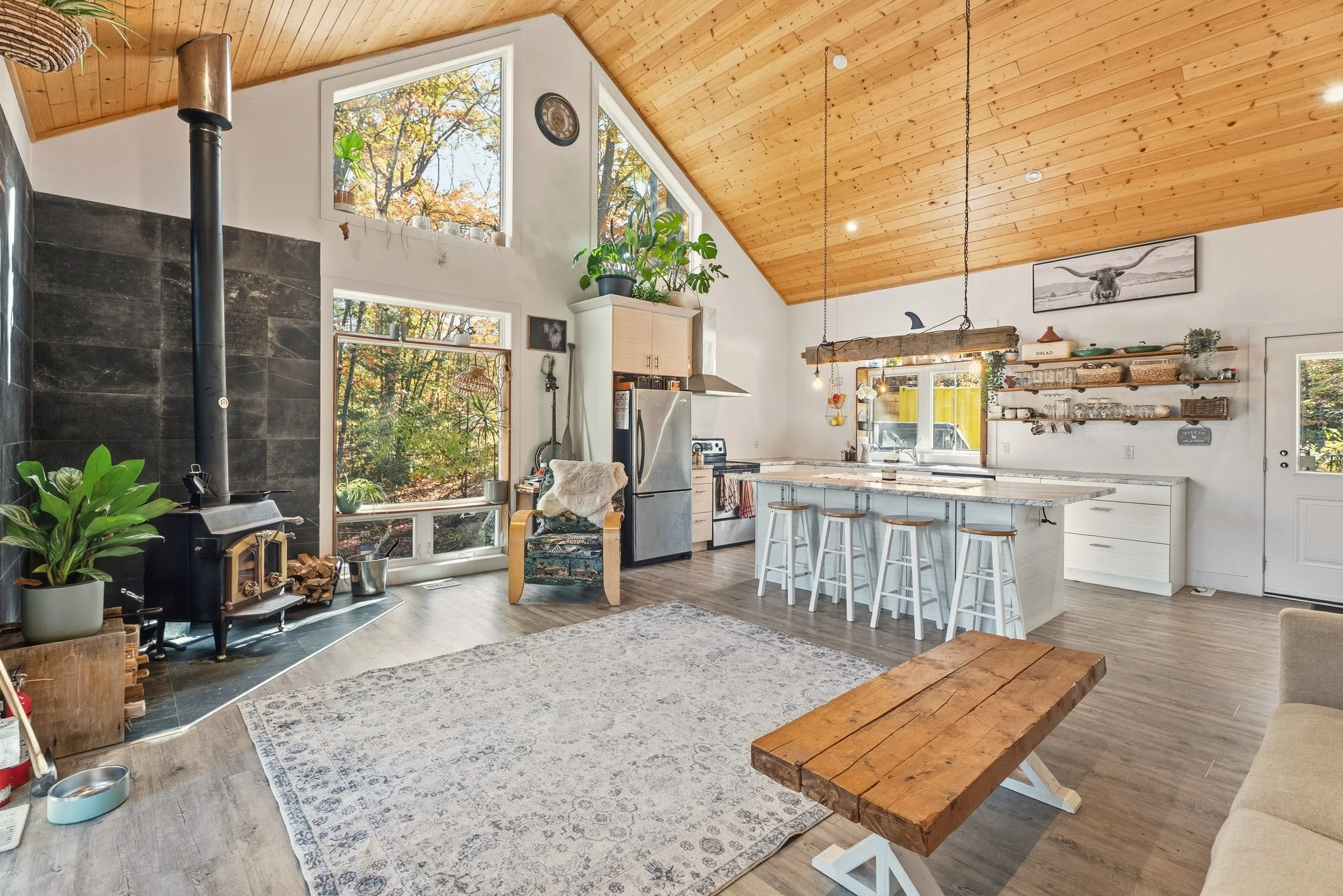 Open-concept kitchen and living room with wood vaulted ceiling, large windows, a wood stove, a rug, a wooden coffee table, and a kitchen island with bar stools.