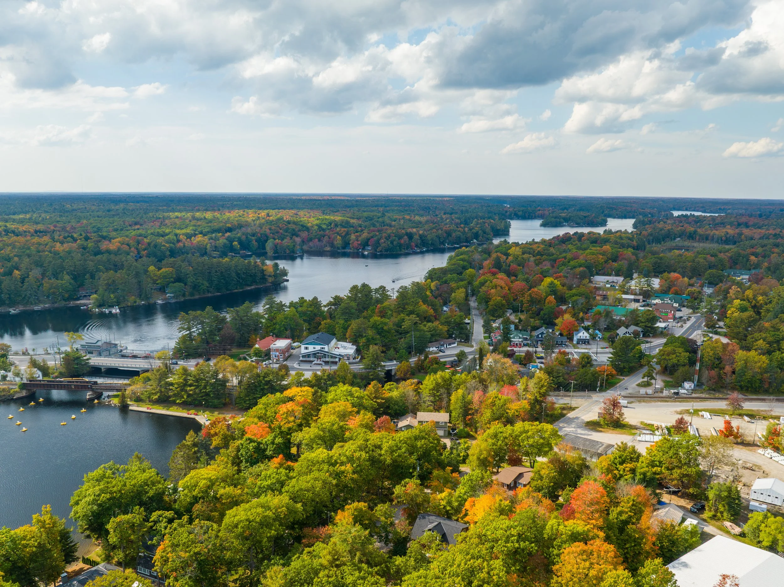 Aerial view of a town surrounded by trees with colorful fall foliage, a river running through the area, and a partly cloudy sky overhead.