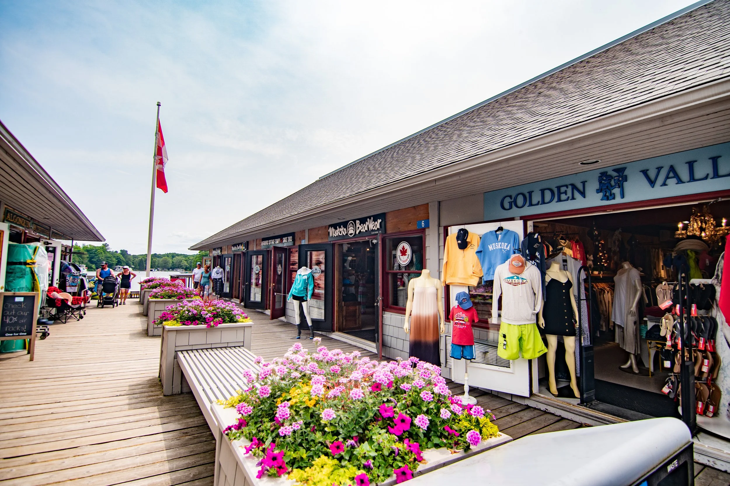 View of a shopping area on a boardwalk with shops selling clothing and accessories, colorful flowers in planters, people walking and shopping, and an American flag flying overhead on a partly cloudy day.