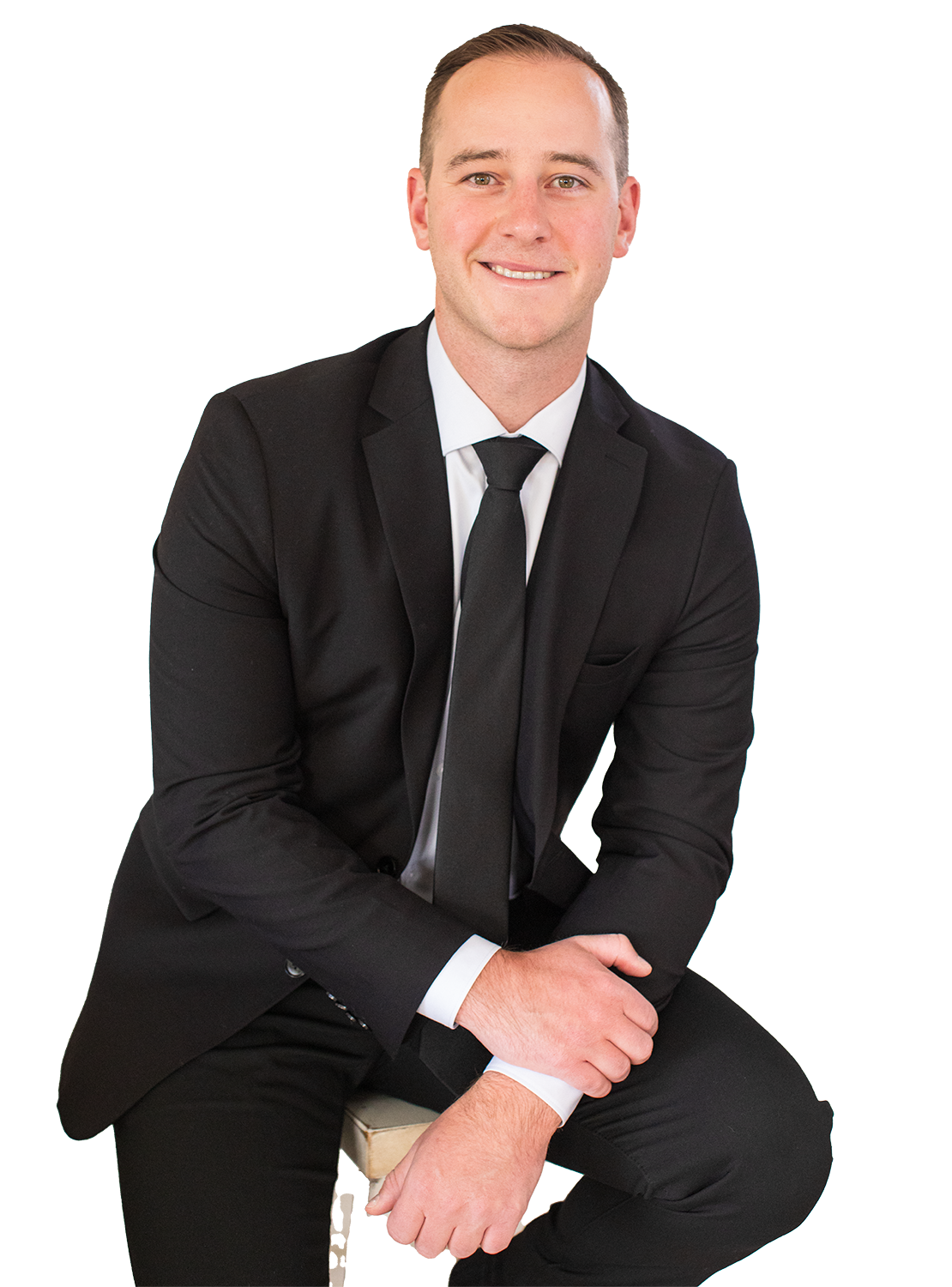 A man in a black suit, white shirt, and black tie sitting on a stool, smiling for a portrait against a plain background.