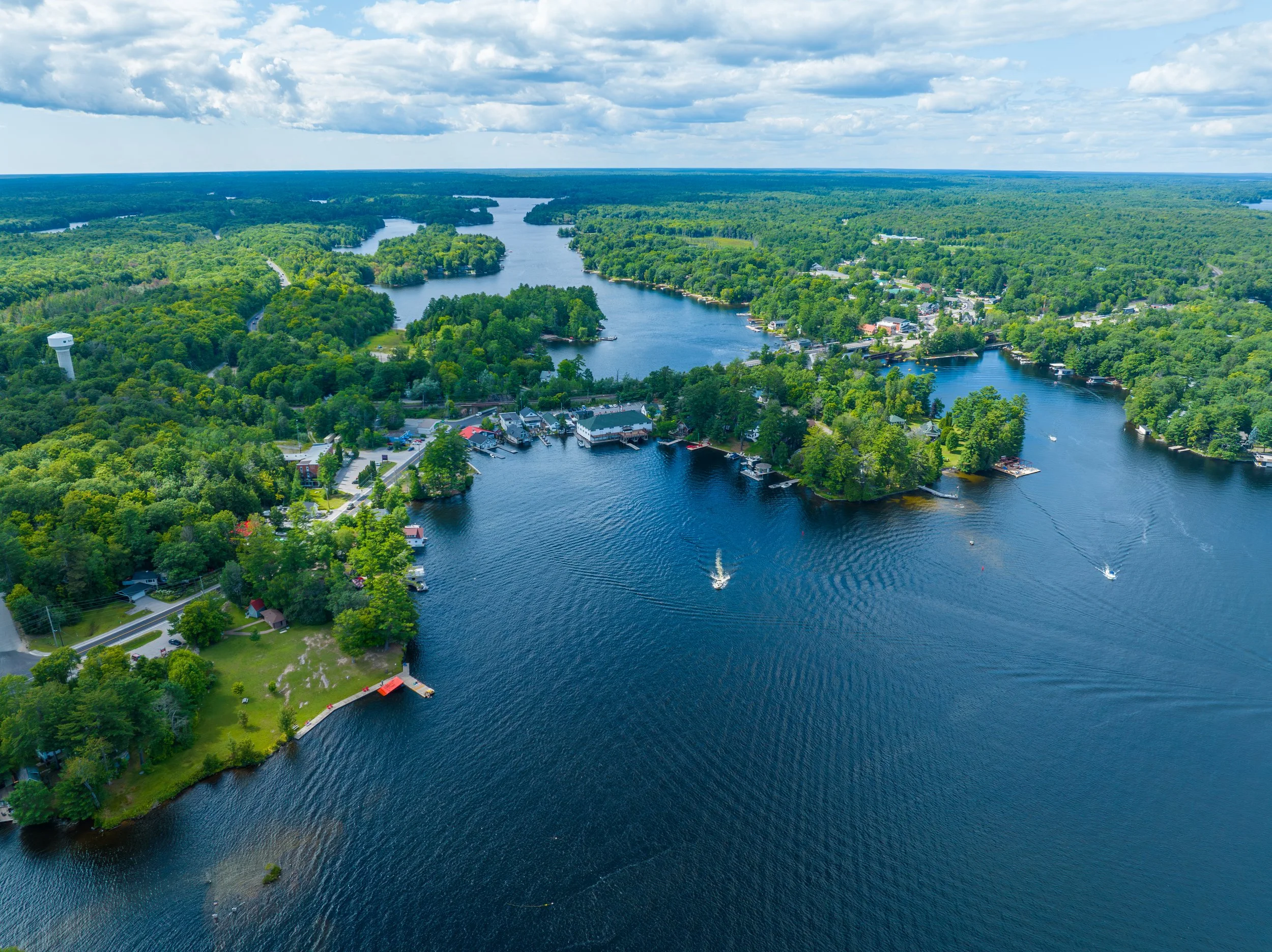 Aerial view of a river flowing through a lush green landscape with boats on the water, houses along the shoreline, and a partly cloudy sky overhead.