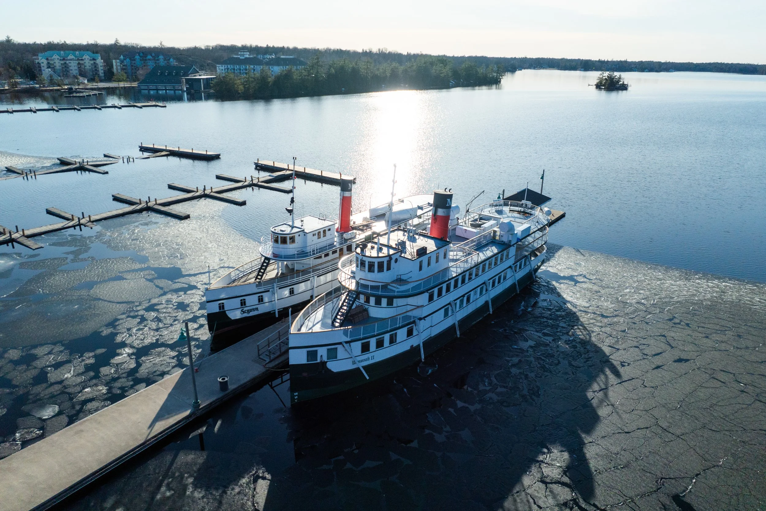 Two vintage paddle steamers docked at a pier on a partially frozen lake, with a backdrop of residential buildings and trees under a clear sky.