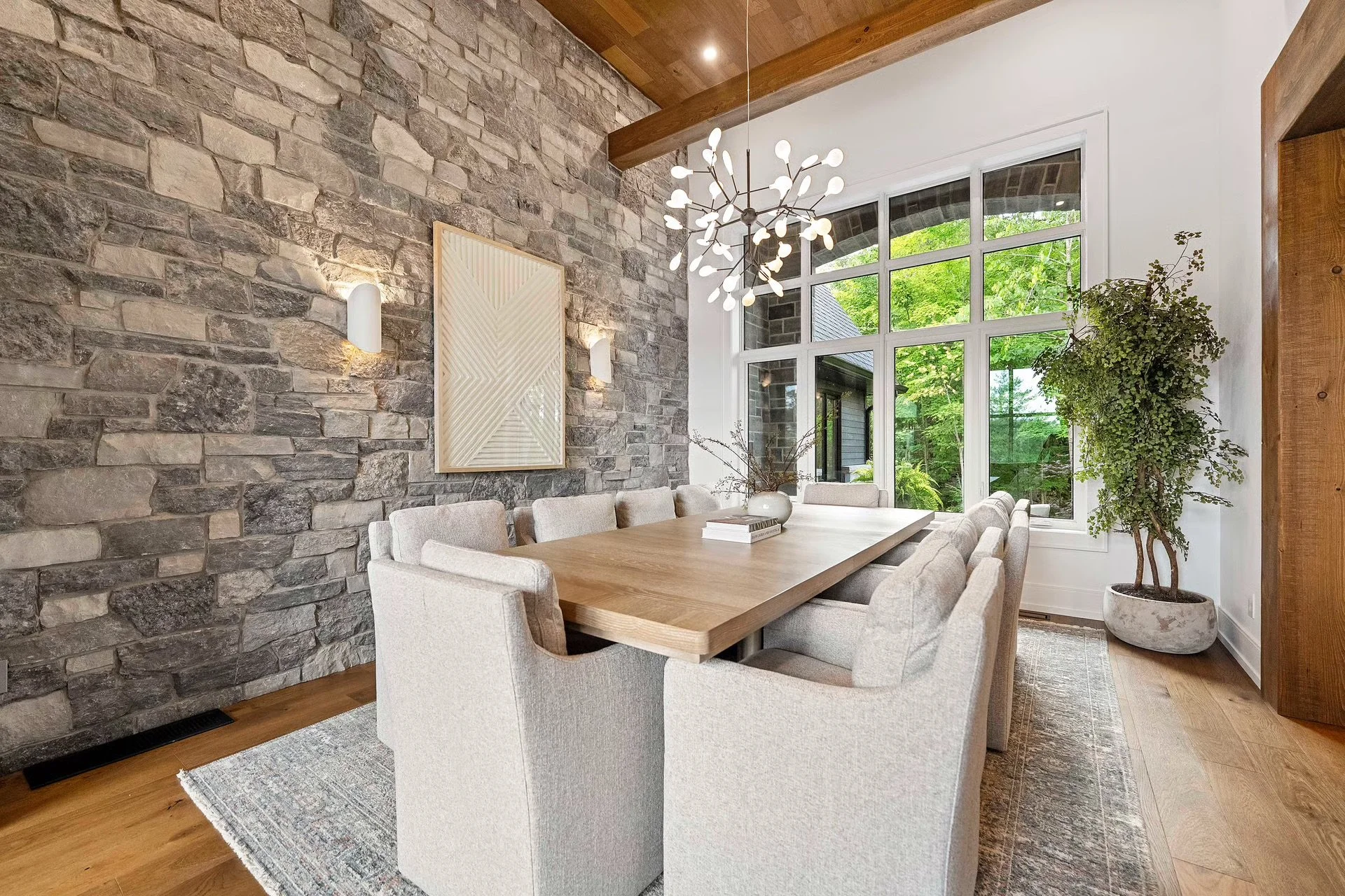 A modern dining room with a long wooden table surrounded by beige upholstered chairs. The room features a gray stone accent wall with wall-mounted lights, a large window with a view of greenery outside, a decorative chandelier hanging from a wooden ceiling, and a potted plant near the window.
