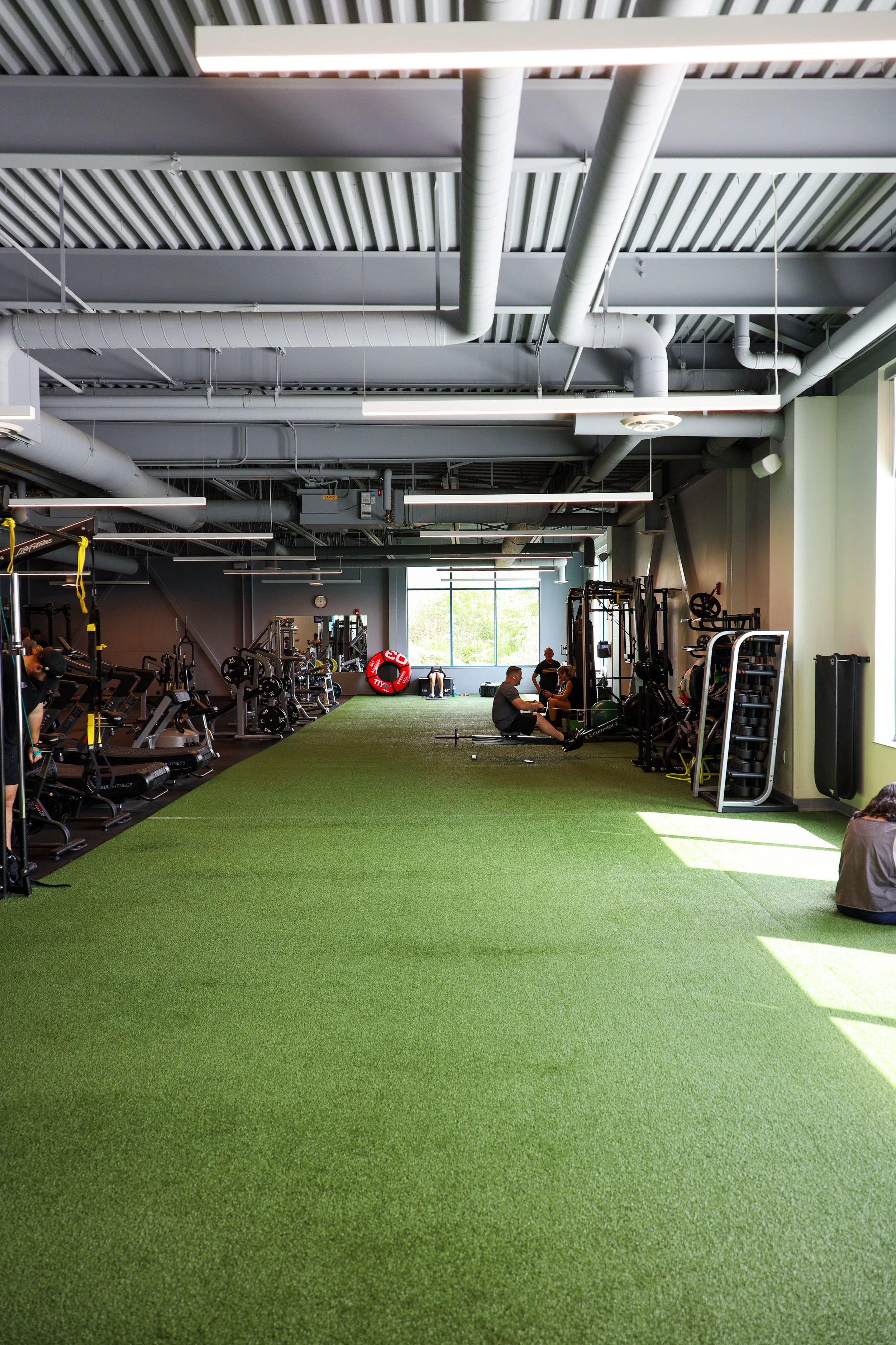 Interior of a gym with green turf flooring, weight machines, and people exercising near large windows.