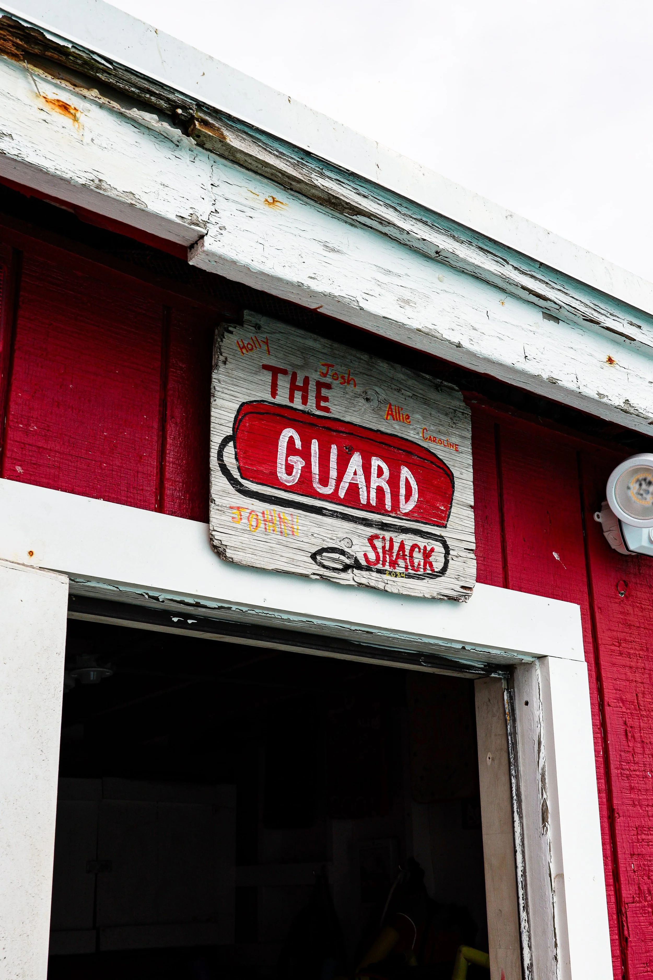Wooden sign with painted text reading "The Guard Shack" above the entrance of a small building, with names "Holly, Josh, Allie, Caroline, John" written around it, red and white painted exterior, and a light fixture to the right.