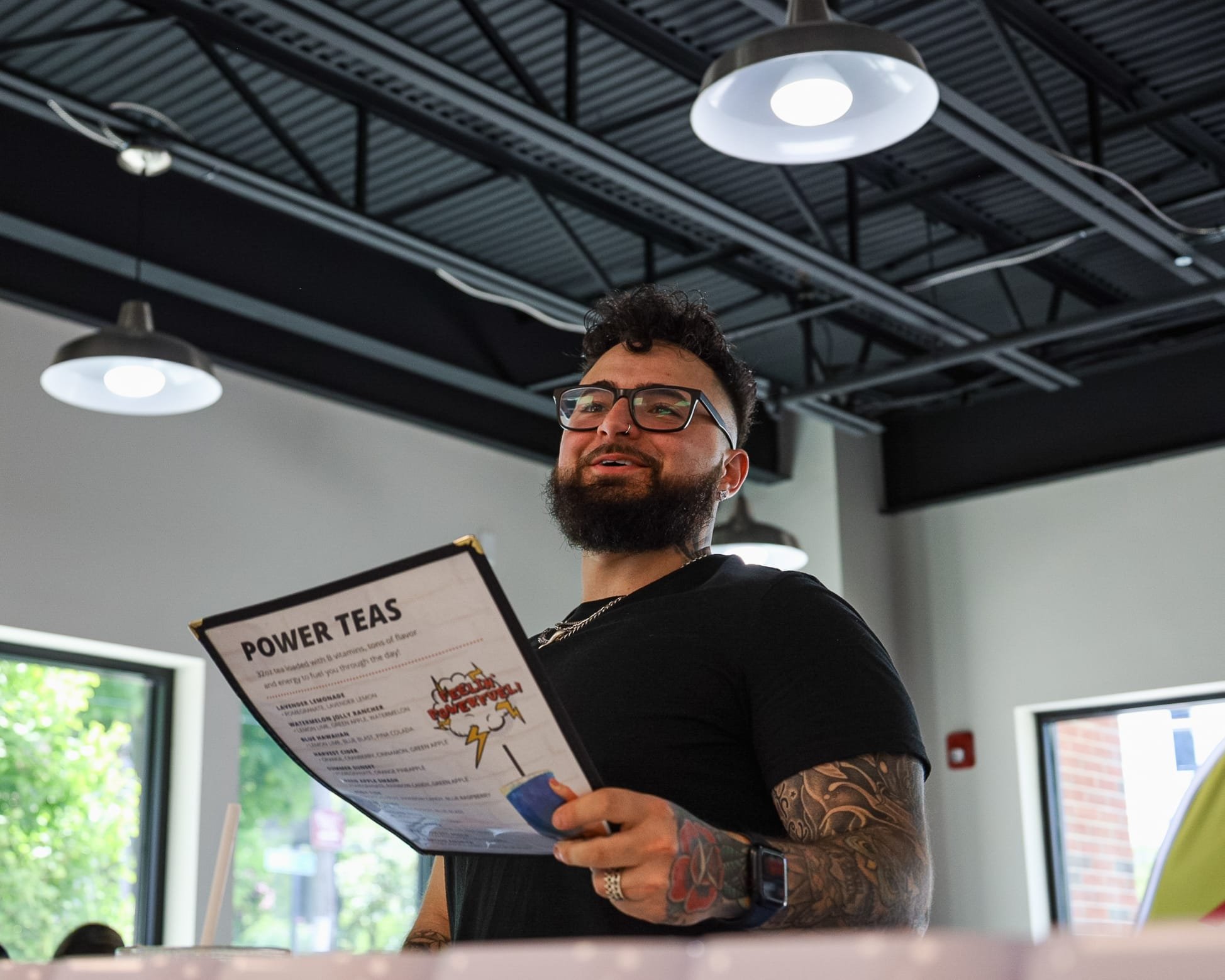 A man with glasses, tattoos, and a beard holds a menu titled "Power Teas" inside a modern cafe with large windows and industrial ceiling lights.