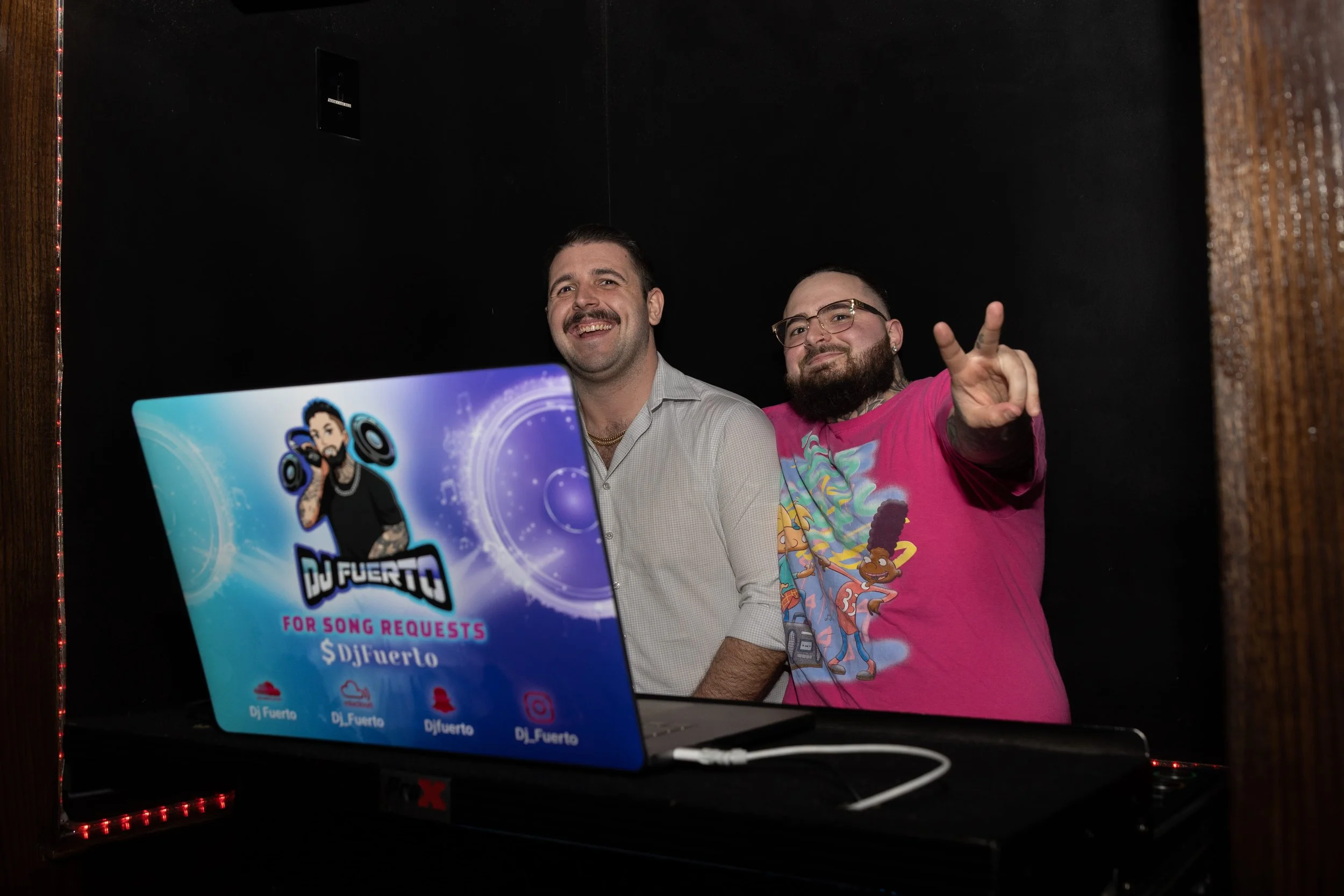 Two men standing behind a DJ booth with a laptop displaying a DJ logo, one in a light-colored shirt and the other in a pink T-shirt making a peace sign, both smiling in a dark room.