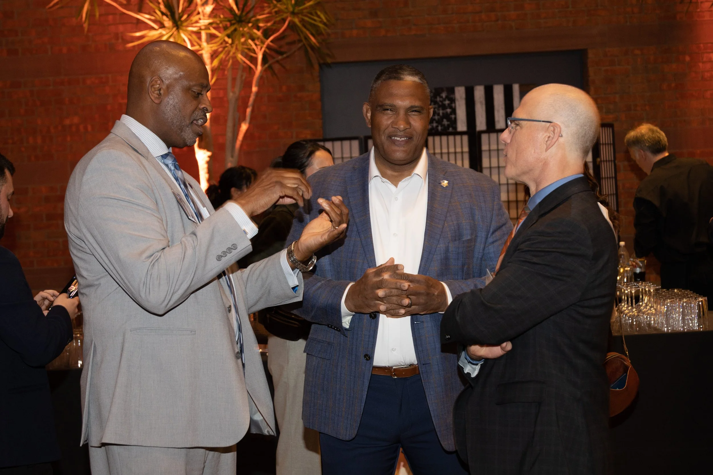 Three men in suits engaged in conversation at a professional event, with a brick wall and a small tree in the background.