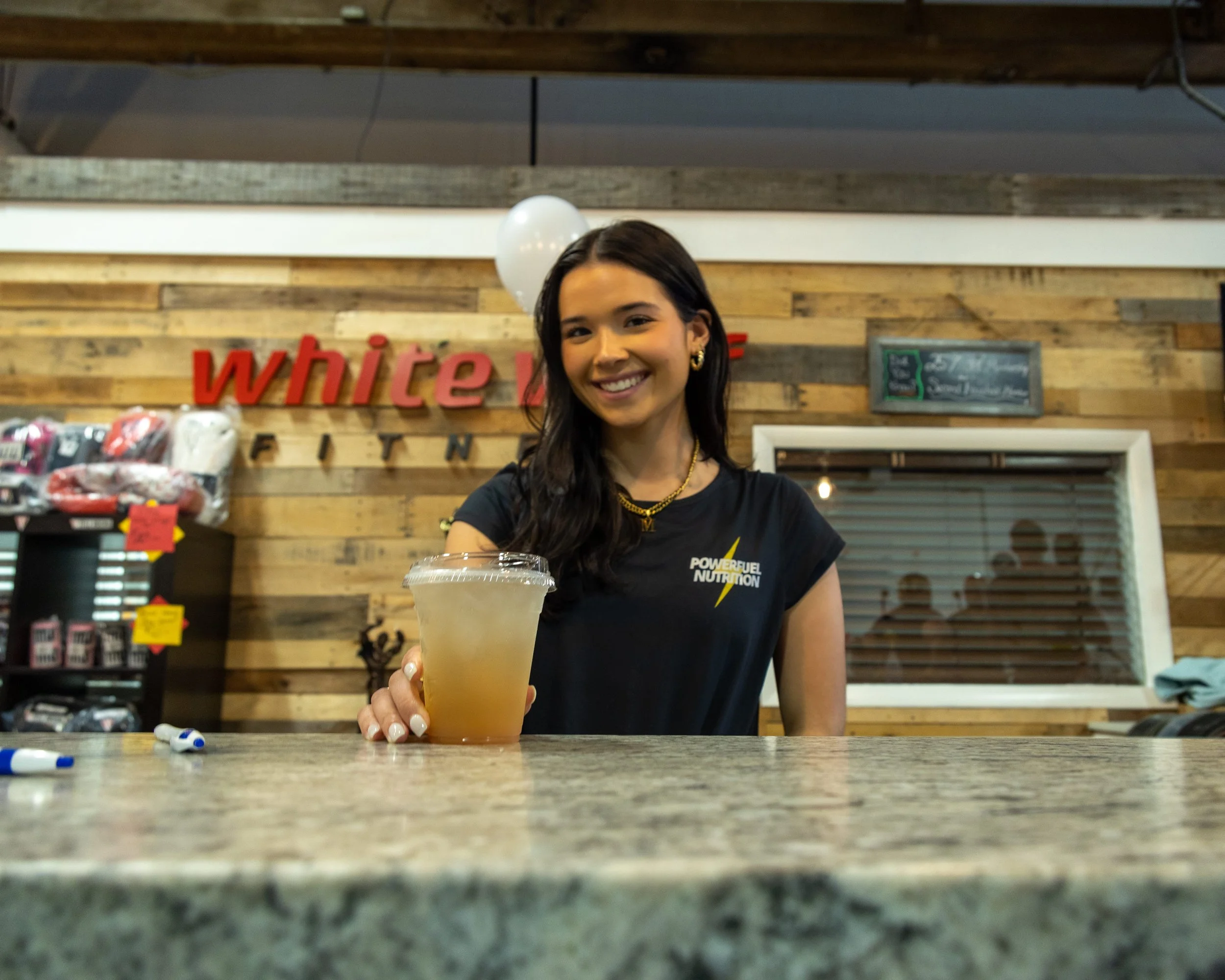 A smiling woman behind a counter holding a plastic cup with a drink inside at a fitness center with a wooden wall background and a sign reading 'whey' in red letters.