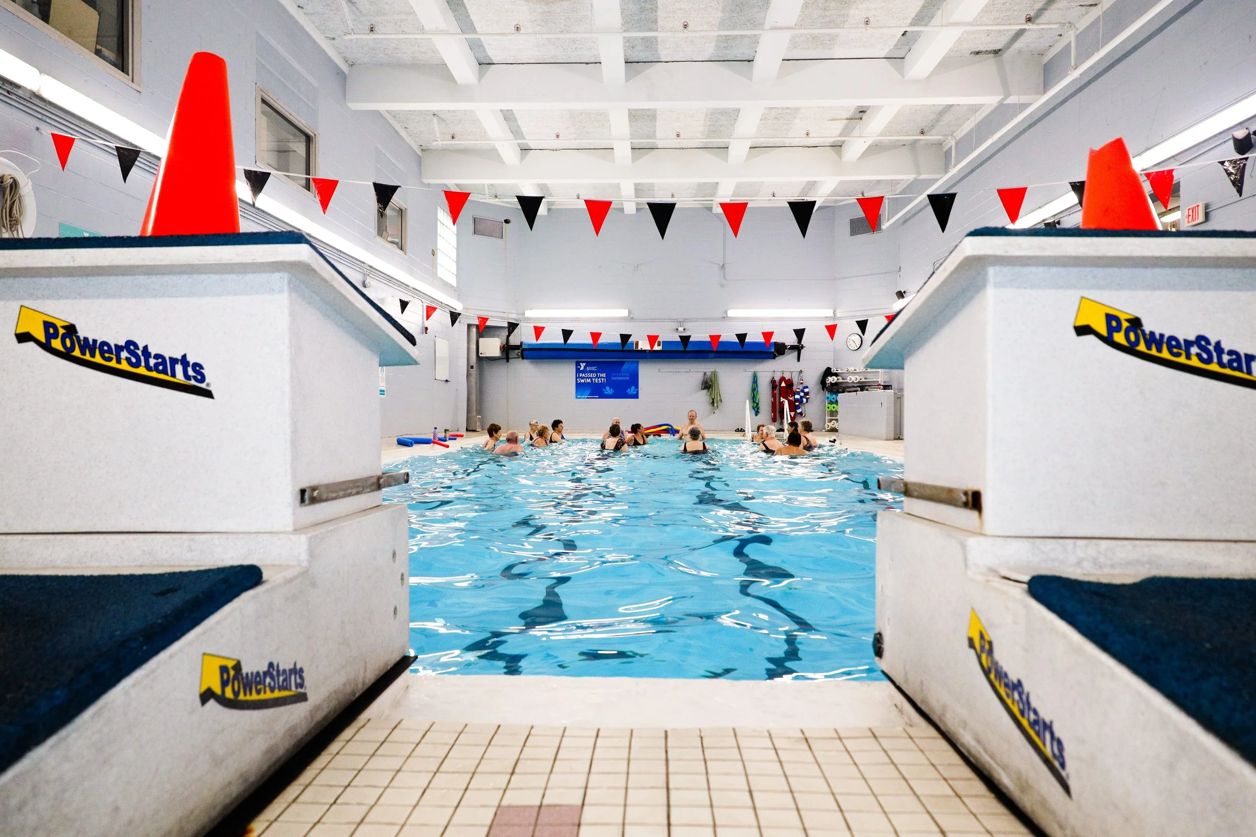 Indoor swimming pool with a group of people participating in a swim class or exercise session. Pool is seen from the shallow end, with starting blocks marked 'PowerStarts' in the foreground, and red and black triangular flags hanging above. The background features a coach or instructor giving guidance, and the pool area is adorned with rescue equipment and colorful towels.