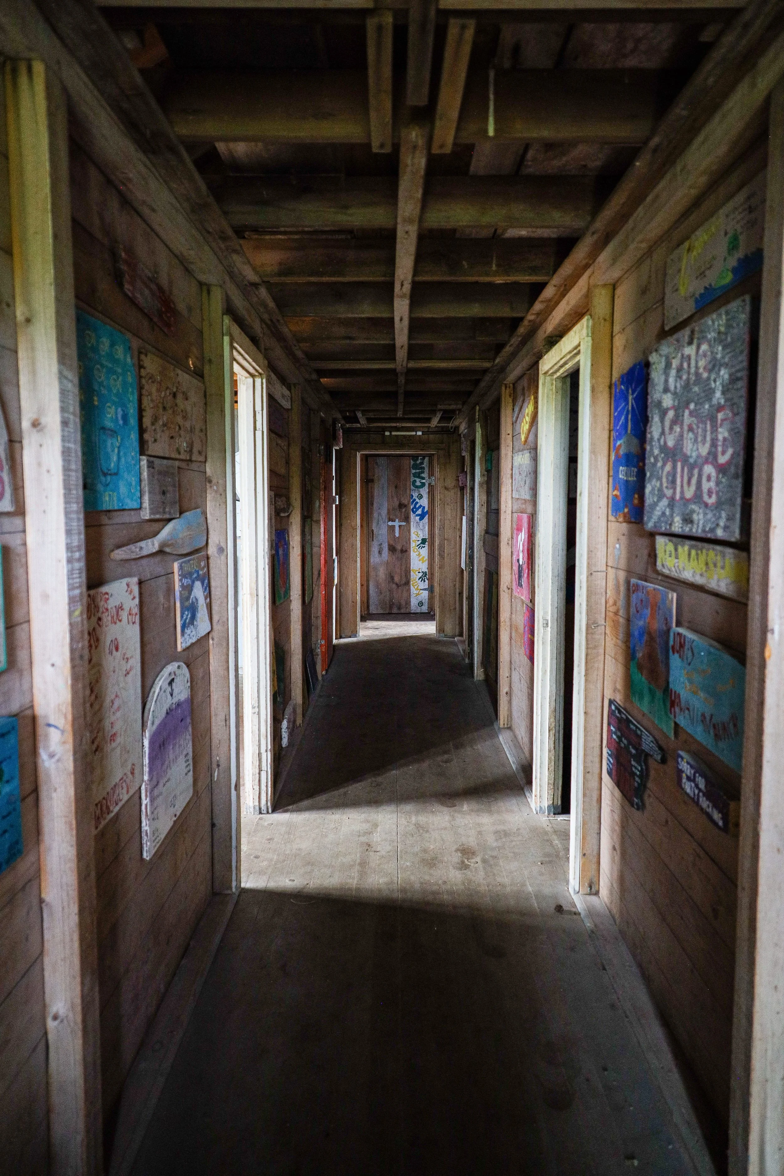 A narrow wooden hallway inside a rustic building, with colorful paintings and drawings displayed on the walls.