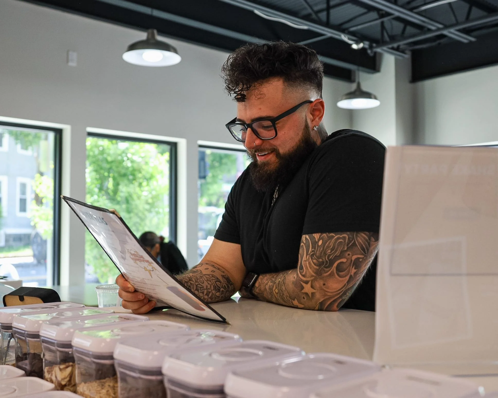 A man with tattooed arms, glasses, and a beard smiling while looking at a menu in a modern cafe or bakery with large windows and bright natural light.