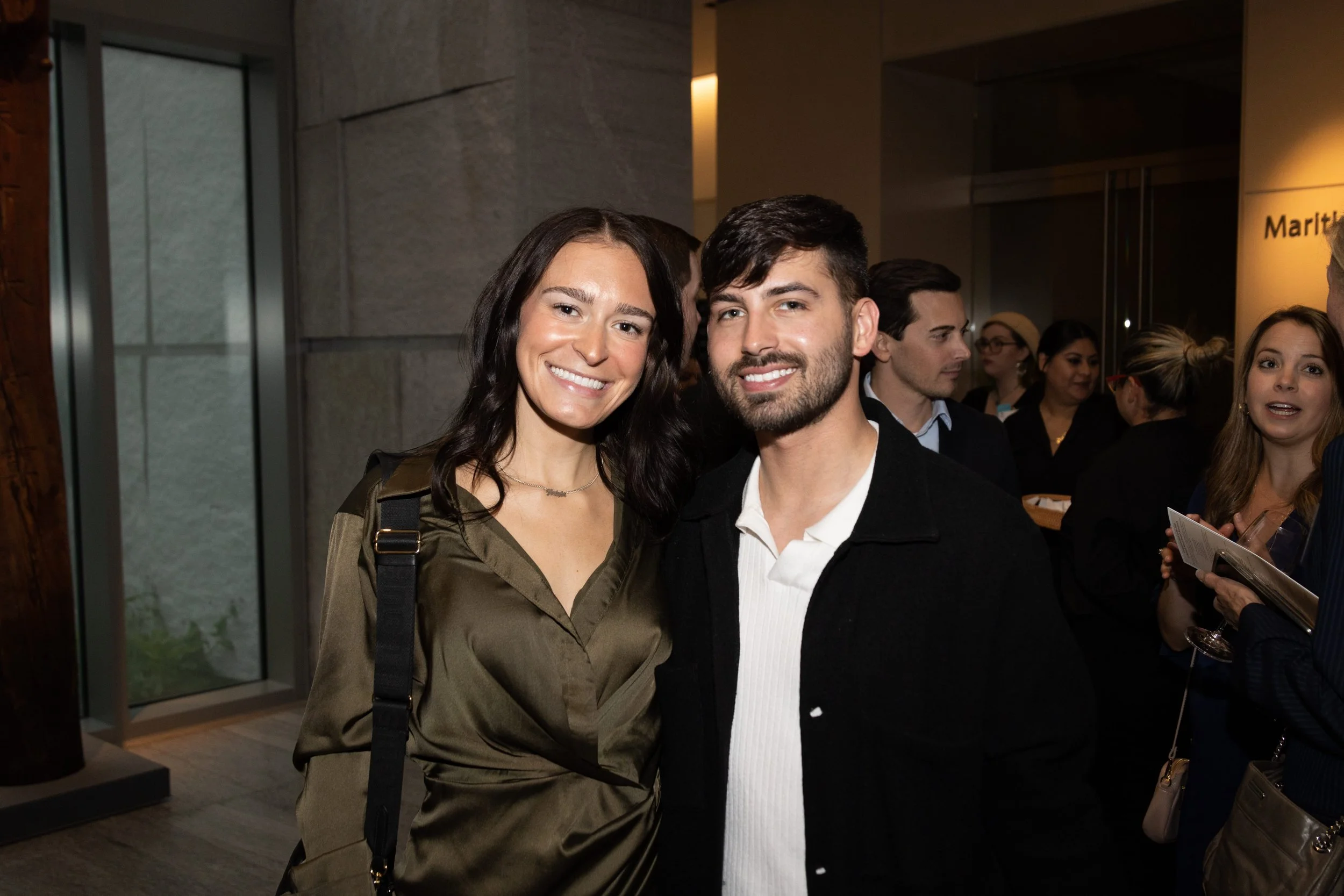 A smiling woman with dark hair and a man with dark hair and facial hair standing close together at an indoor event, surrounded by other people.